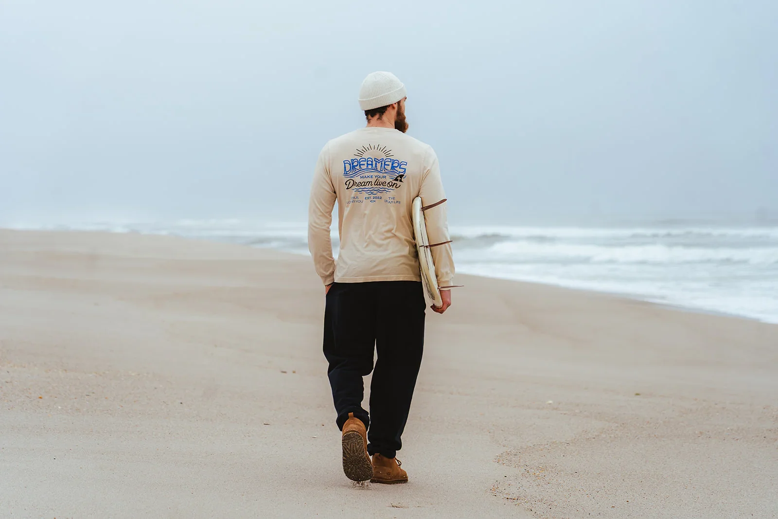 Man walking on the beach holding a surfboard, wearing a beige sweatshirt with 'Dreamers' printed on the back, black pants, brown shoes, and a white beanie.