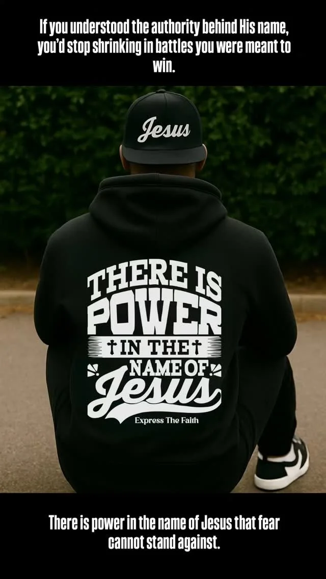 A person kneeling outdoors, wearing a black hoodie with the phrase "There is power in the name of Jesus" on the back and a black cap with the word "Jesus" written on it. The background is blurred green foliage.