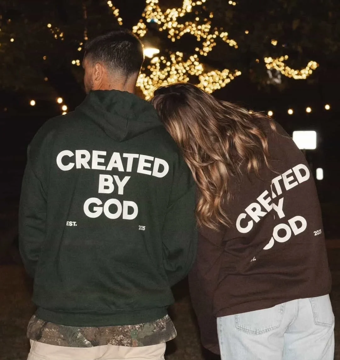 Two people standing outdoors at night with lights in the background, wearing hoodies that say 'Created by God' on the back.