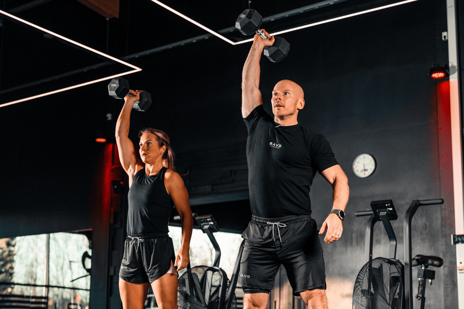 Two people lifting dumbbells in a gym, a woman on the left and a man on the right, both wearing black athletic clothing.