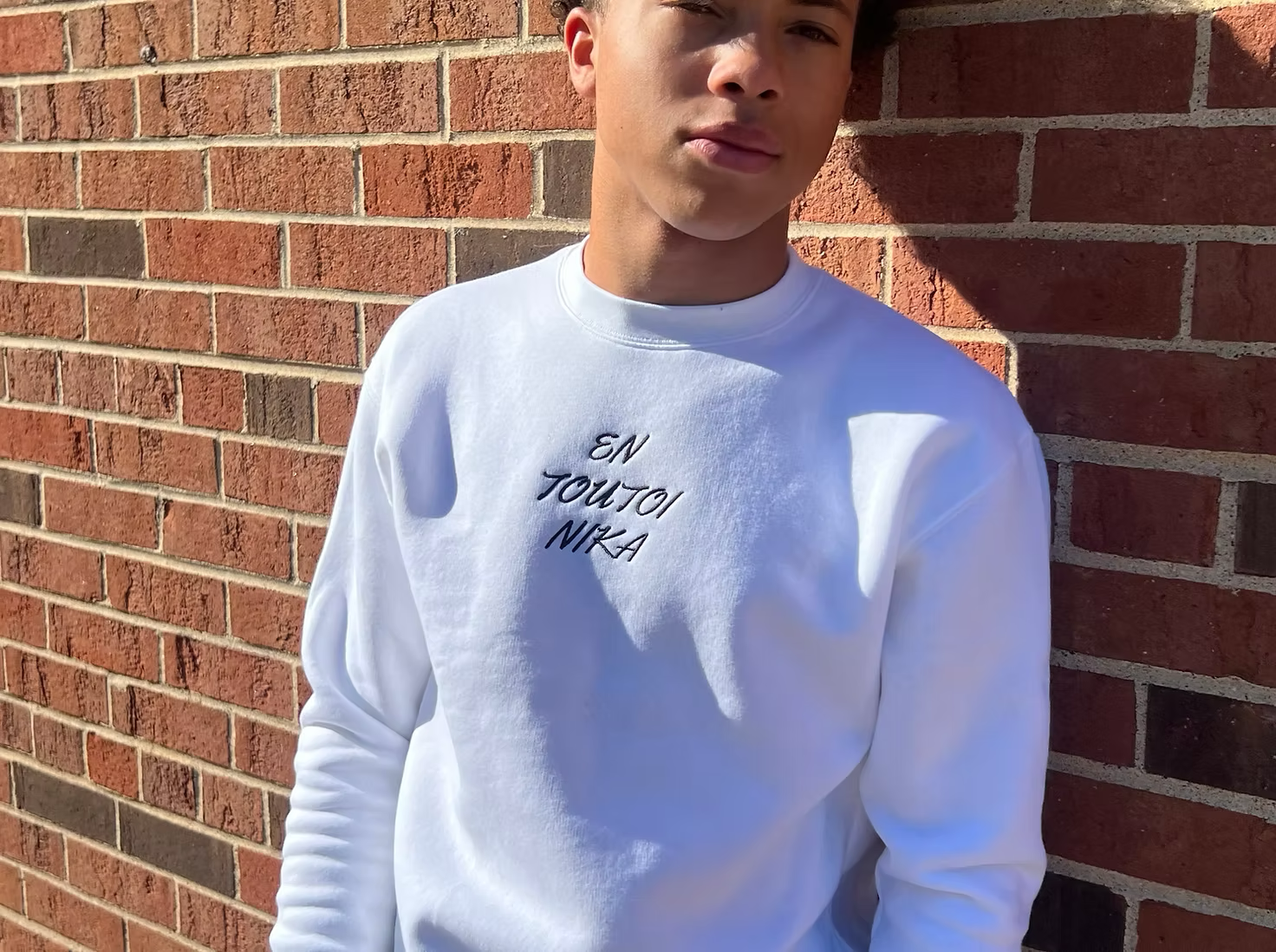 A young man in a white sweatshirt standing against a brick wall, with a partial view of his face visible.