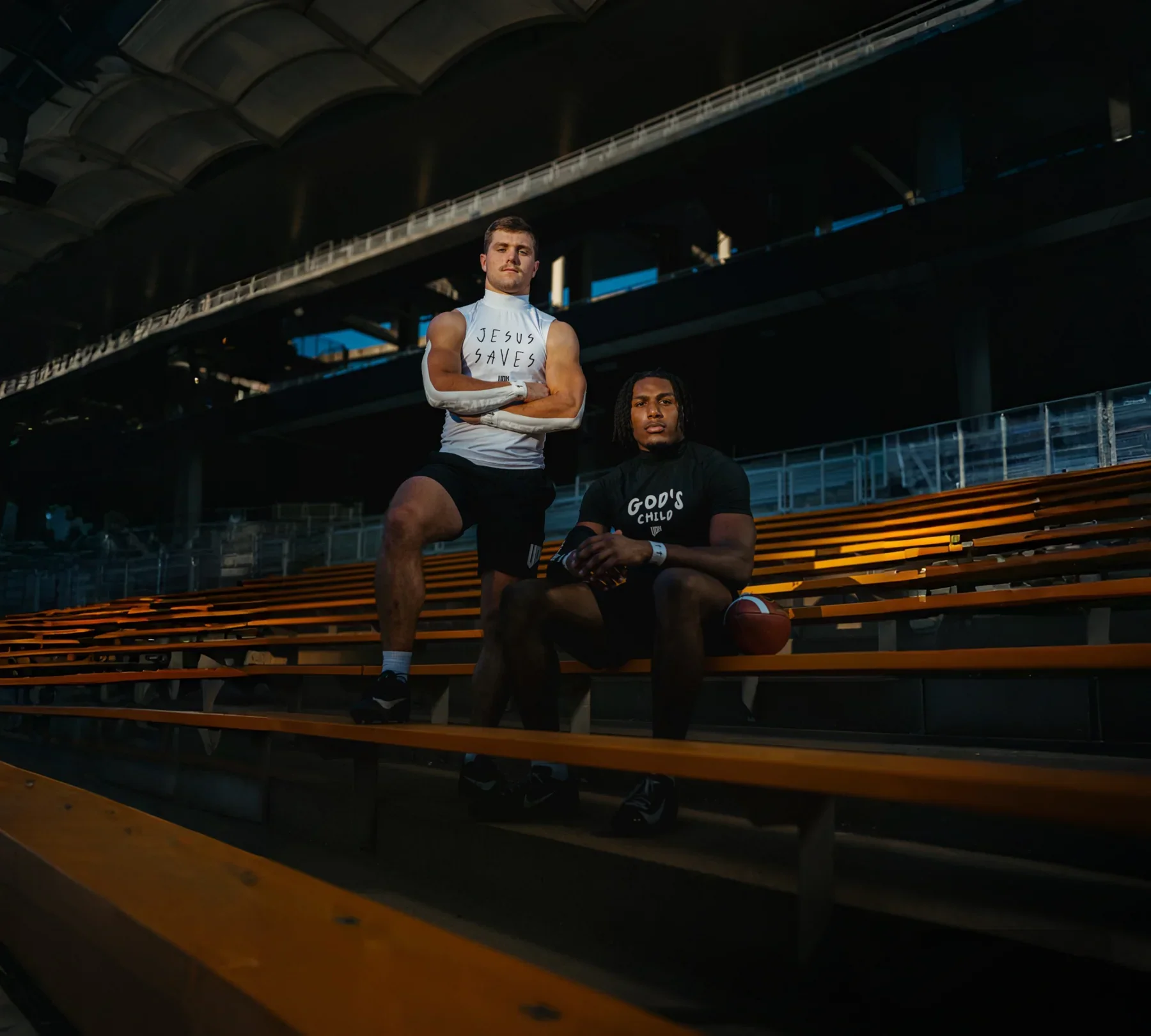 Two football players in athletic gear sitting and standing on empty stadium bleachers during sunset. One player is wearing a white shirt with 'JESUS SAVES' written on it, and the other is wearing a black shirt with 'GOD'S CHILD' written on it, both holding footballs.