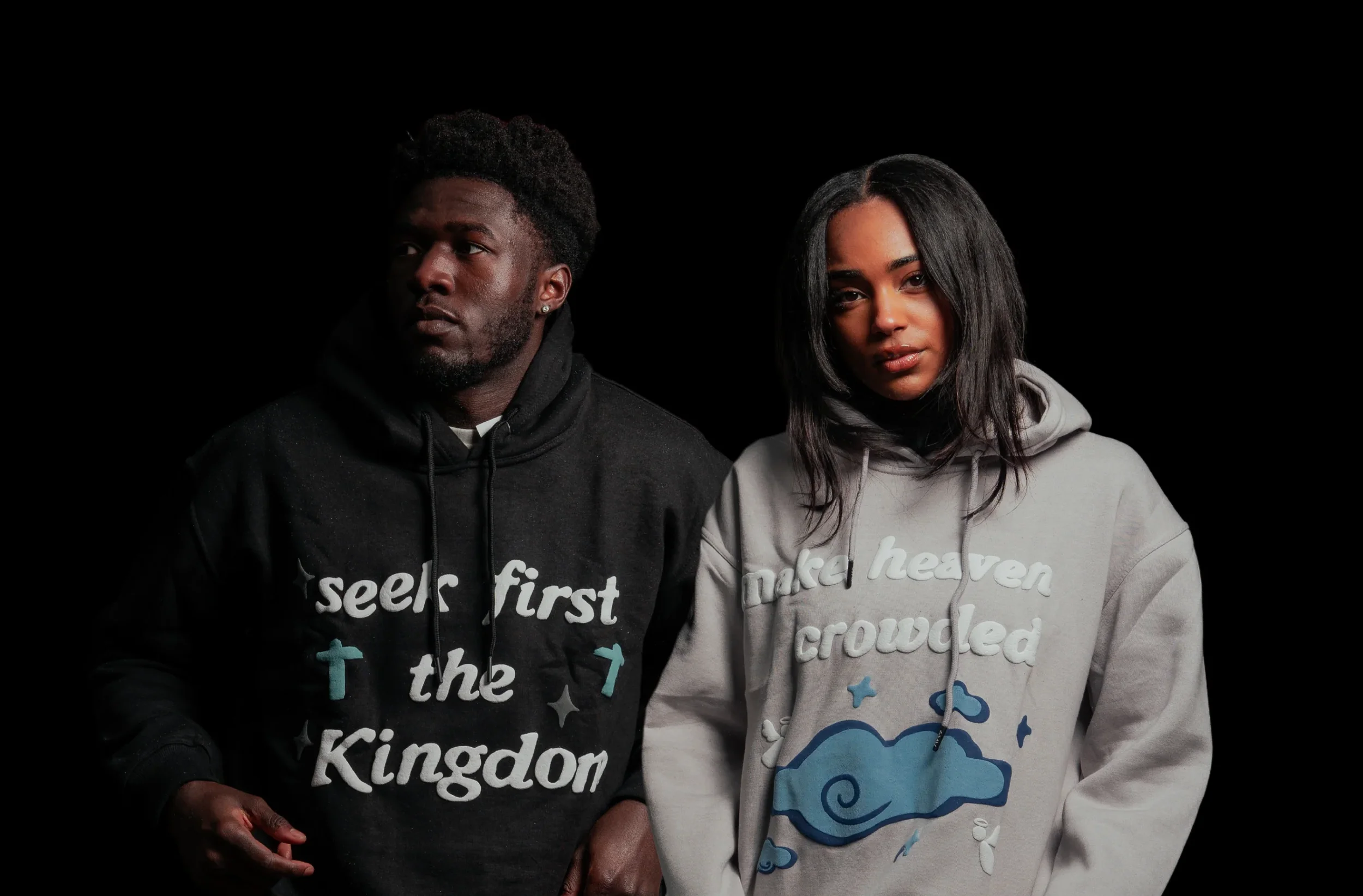 A man and woman standing side by side against a black background, wearing hoodies with religious and inspirational slogans, the man in a black hoodie and the woman in a light gray hoodie.