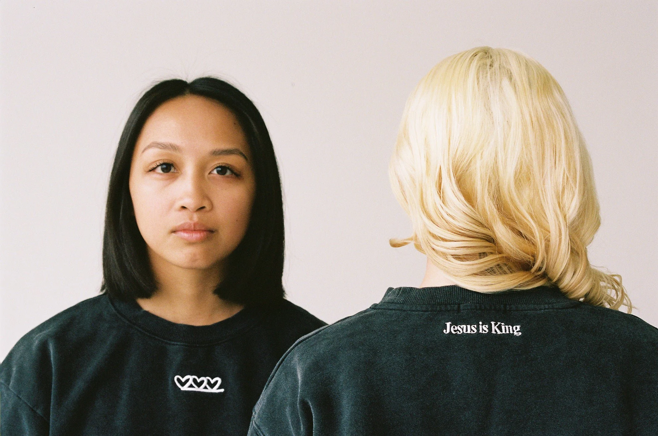 Two women standing side by side, one facing forward and the other turned away. The woman facing forward has black hair and is wearing a black shirt with three hearts on it. The other woman, with blonde hair, is wearing a black shirt with the text 'Jesus is King' on the back.