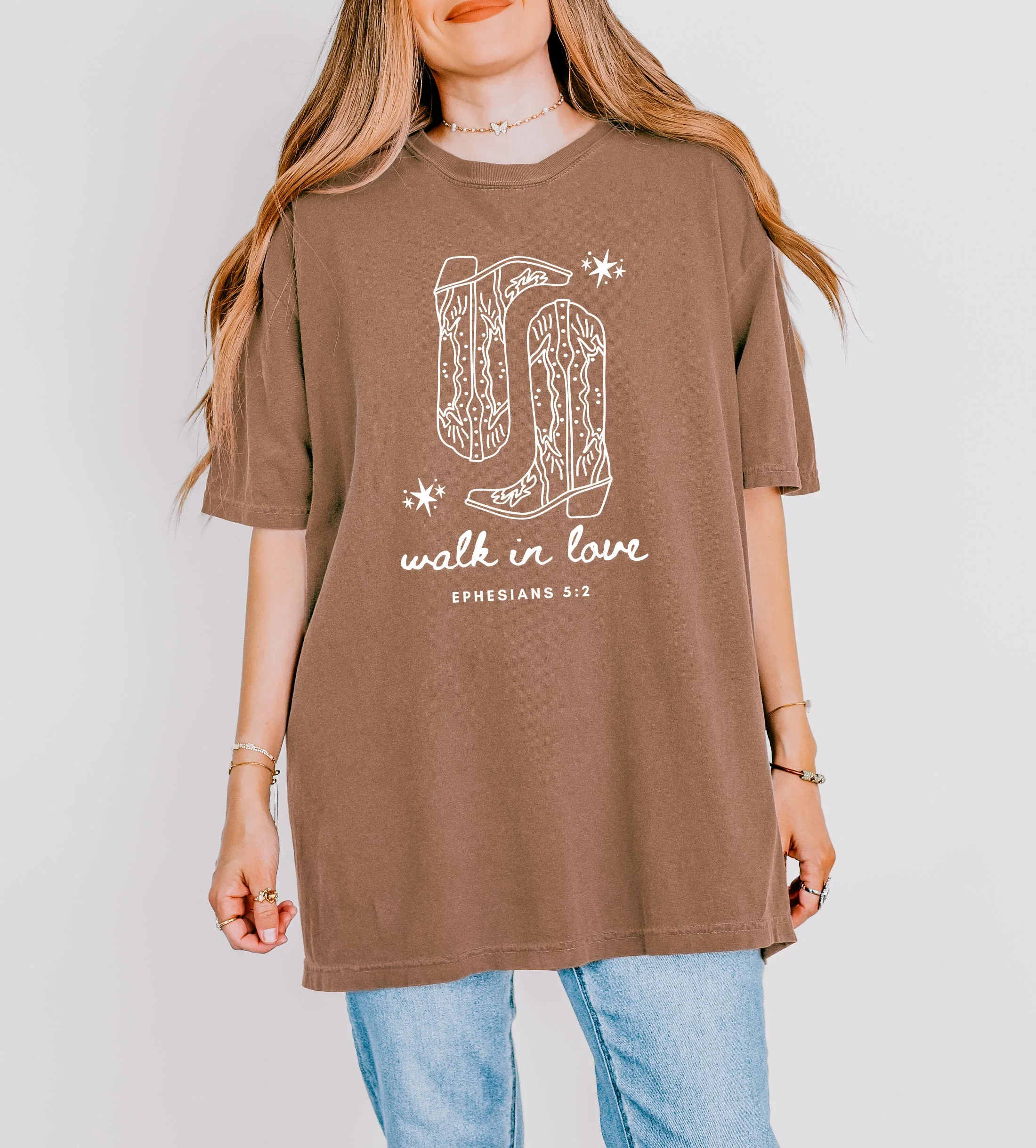 A person wearing a brown t-shirt with a graphic of cowboy boots and a large 'G' and the text 'walk in love' and 'Ephesians 5:2' printed on it. The person has long, light brown hair, and is wearing jewelry including rings, bracelets, and a necklace.