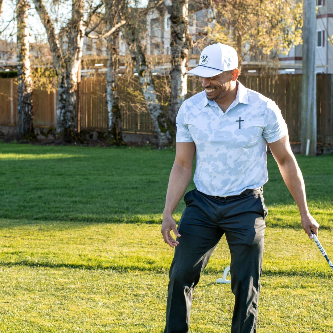 A man wearing a white cap, white patterned polo shirt with a cross, and black pants playing golf on a sunny day in a backyard with grass and trees.