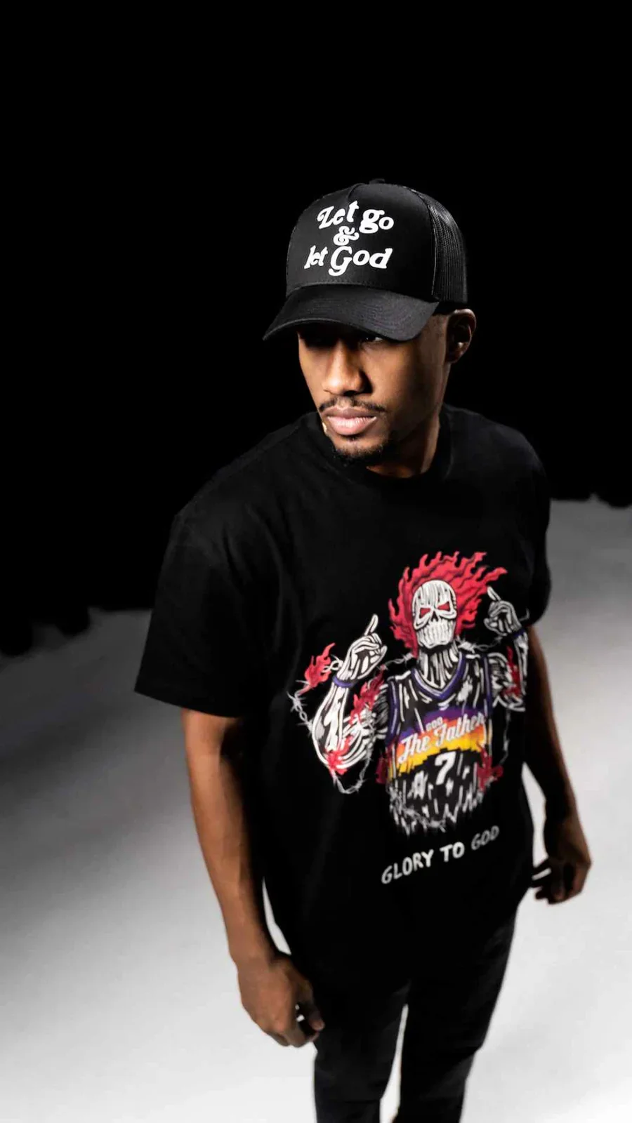 A man wearing a black trucker hat with the phrase 'Let go & let God' and a black T-shirt featuring a graphic of a skeletal figure with fiery hair, raised middle fingers, and the words 'Glory to God.'