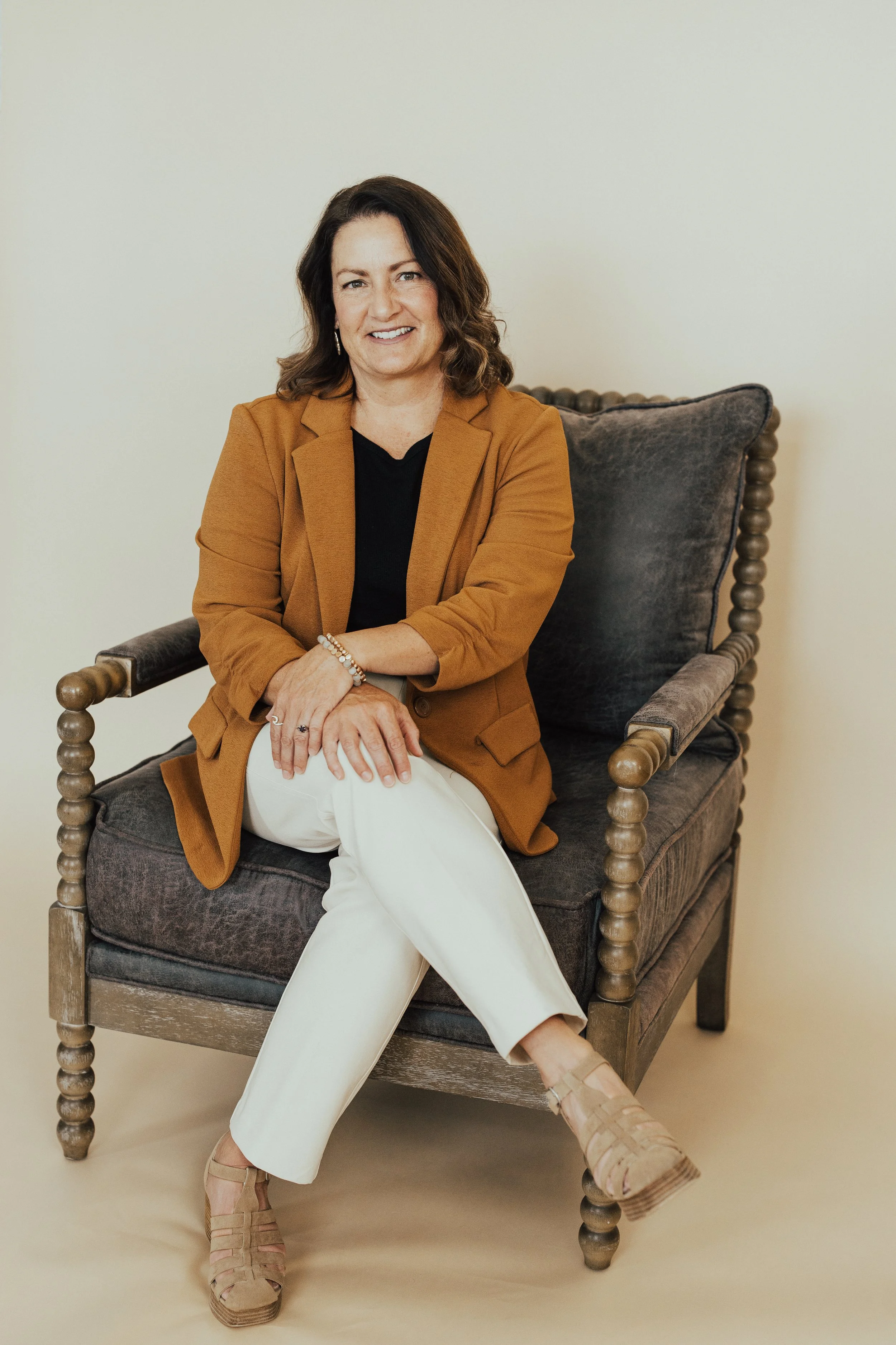 A woman with shoulder-length brown hair, wearing a mustard blazer, black top, white pants, and beige sandals, sitting on a dark upholstered armchair against a plain background, smiling at the camera.