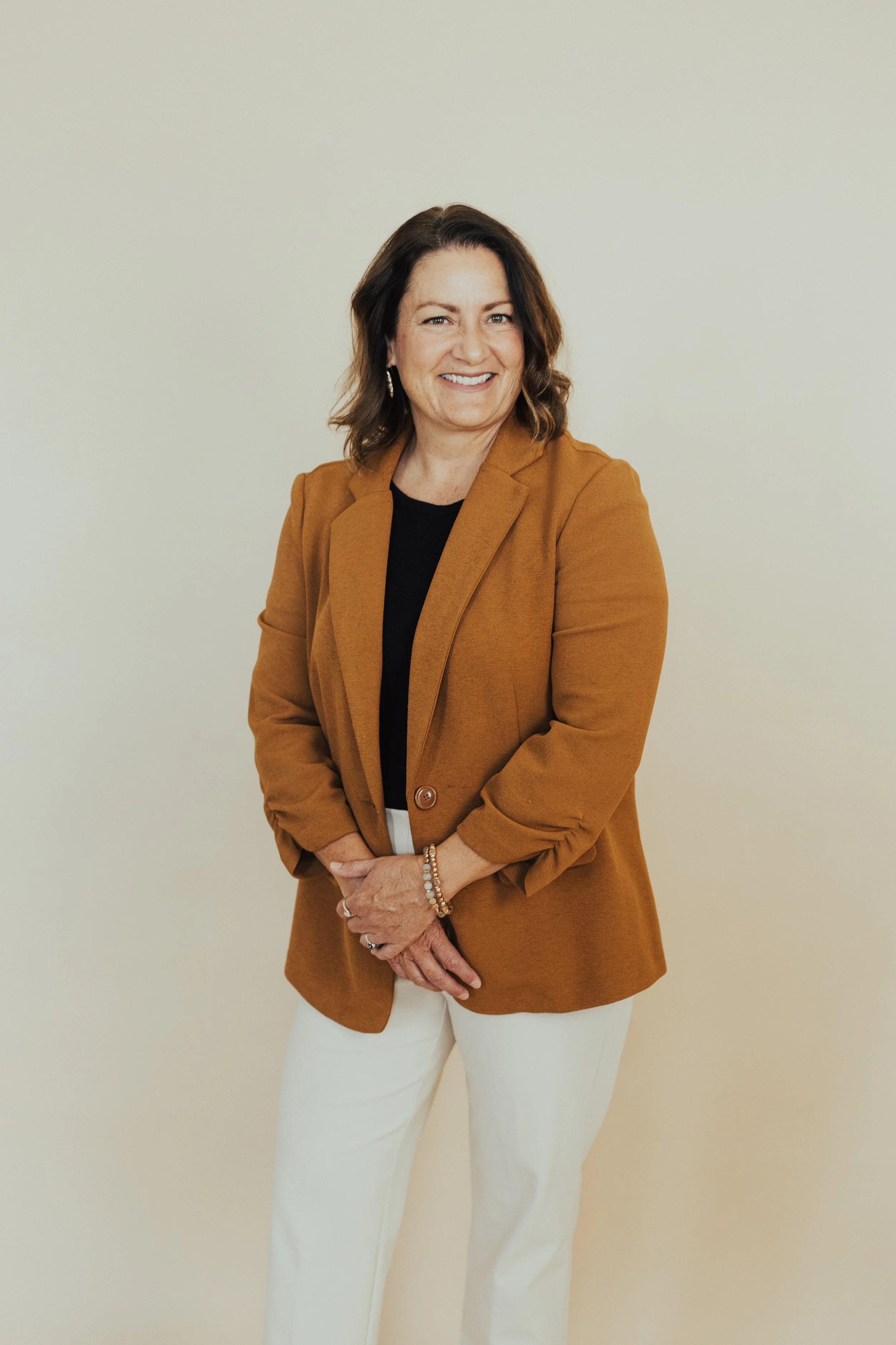 A woman with shoulder-length brown hair, wearing a caramel blazer, black top, and white pants, standing in front of a plain white wall, smiling at the camera.