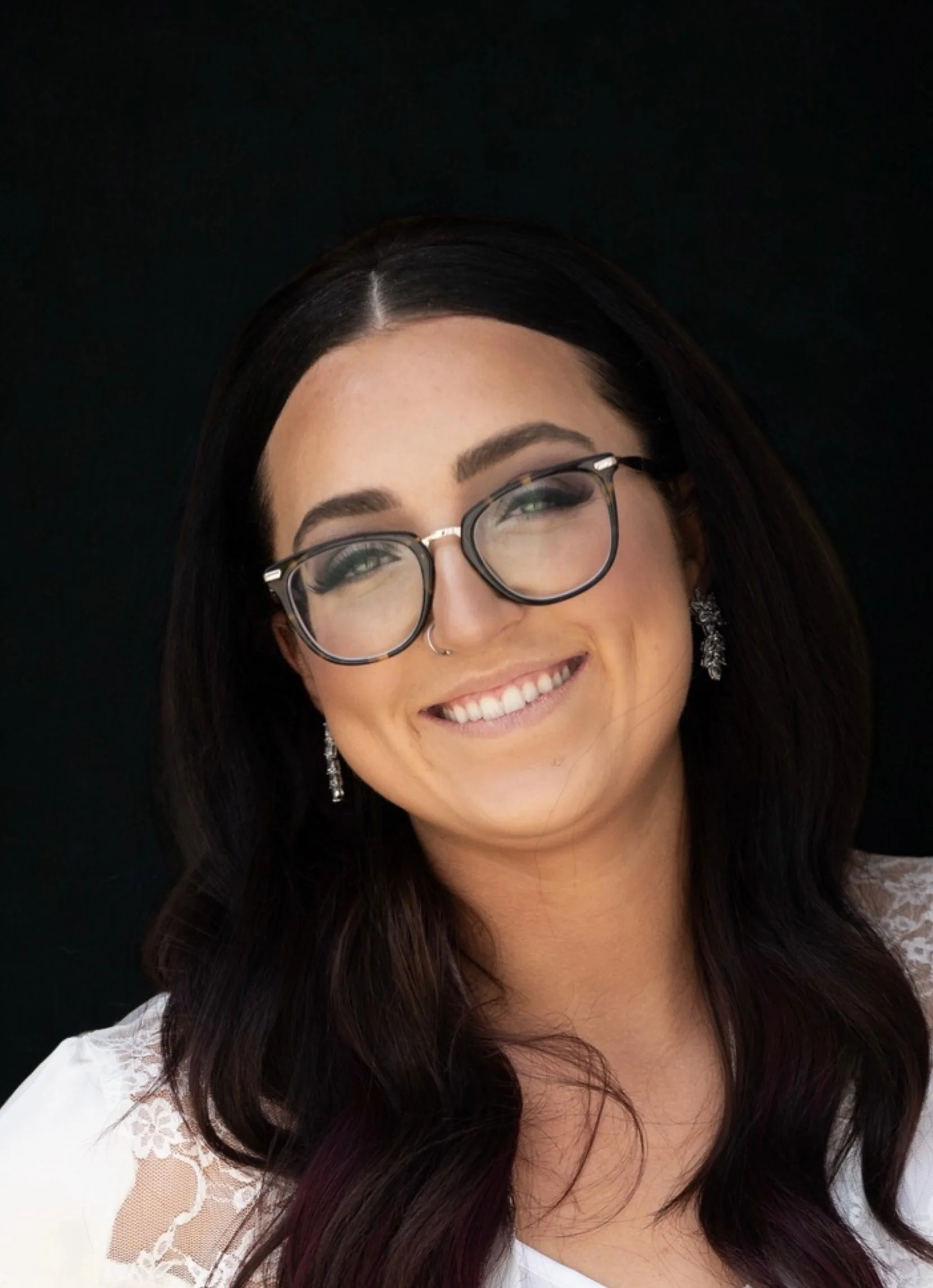 Portrait of a woman smiling, wearing glasses and earrings, with long dark hair and a lace white top, against a dark background.