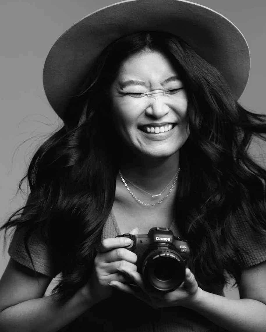 A woman with long dark hair wearing a wide-brimmed hat and jewelry, smiling and winking while holding a Canon camera.