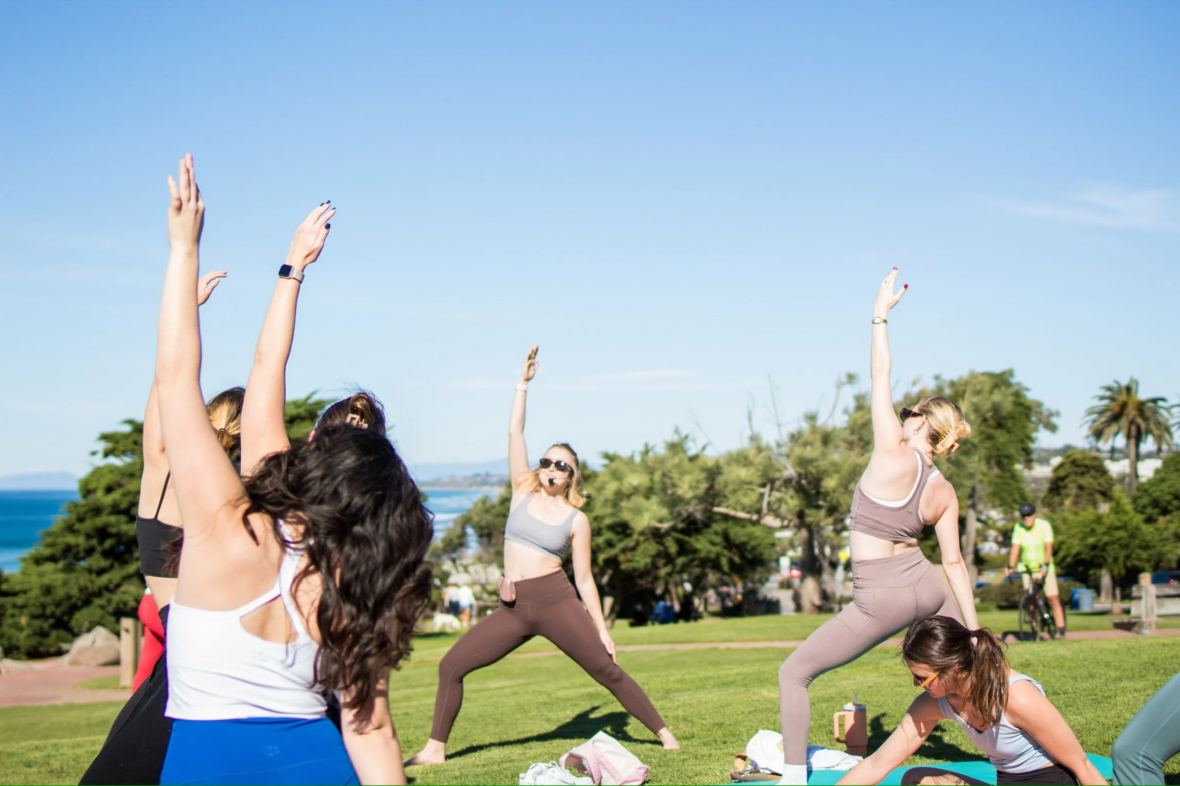 People practicing yoga outdoors on a sunny day in a park with trees and water in the background.