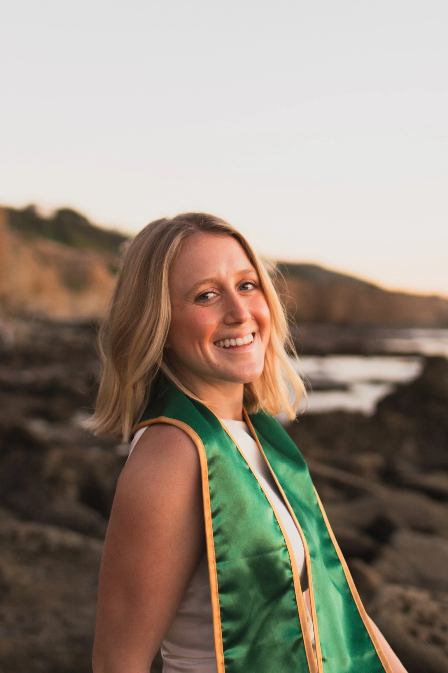 A woman smiling at the camera outdoors during sunset, wearing a green graduation stole over a sleeveless white top, with a rocky coastline and sea in the background.