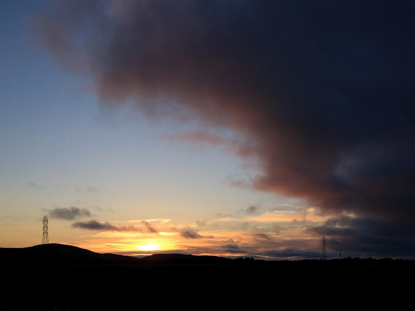 Dark silhouetted hills and two transmission towers beneath a glowing sunset and a heavy band of cloud over the Coyote Hills in Fremont.
