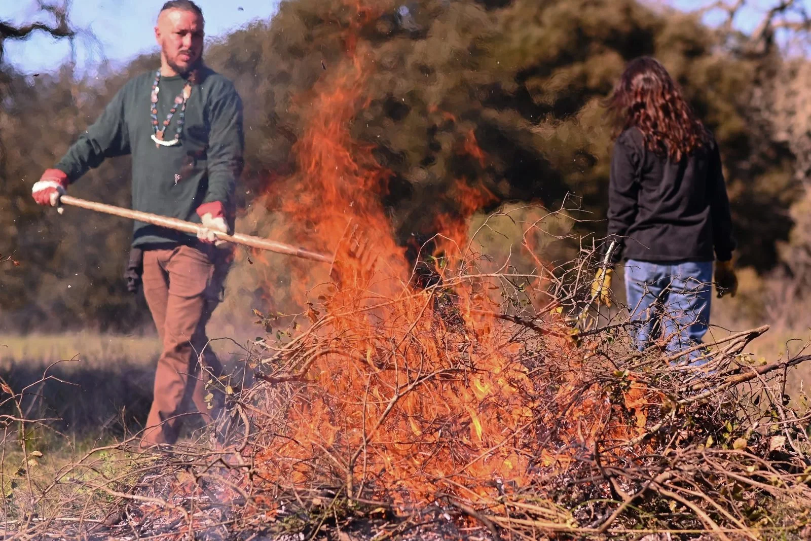 A Muwekma Ohlone tribal member tends a pile burn with a long-handled tool at 'Ootchamin 'Ooyakma