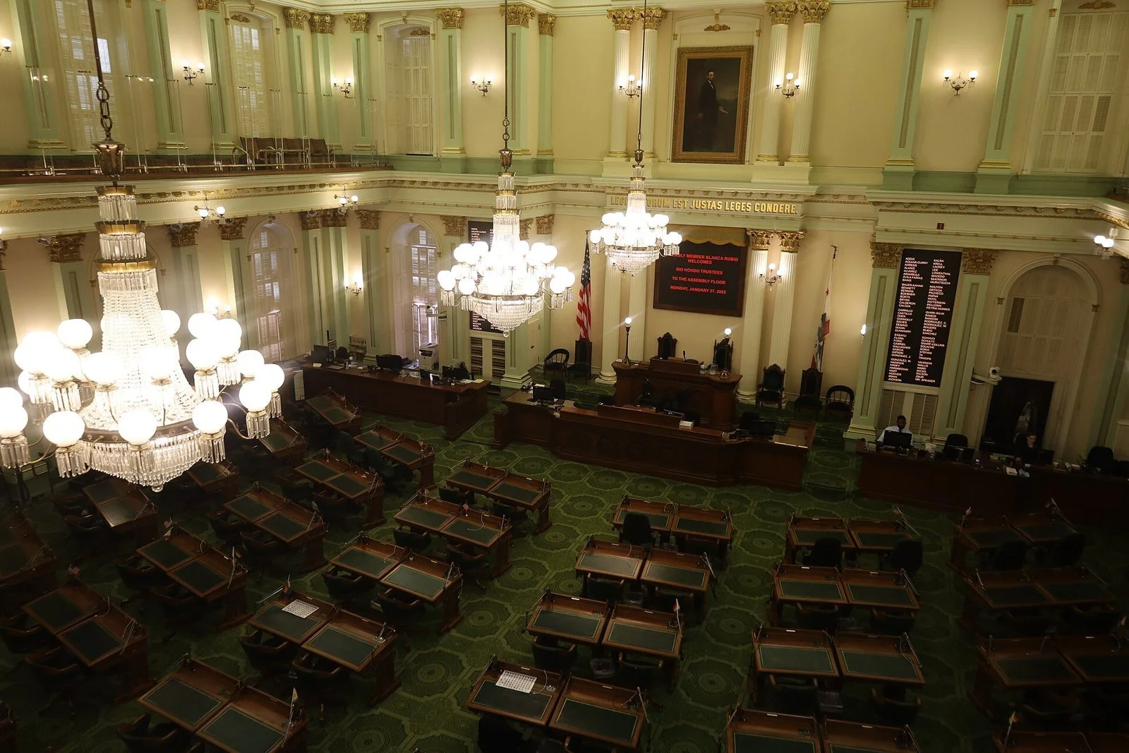 The green-carpeted California Assembly Chamber, with rows of wooden desks, crystal chandeliers and ornate balconies, seen from above.
