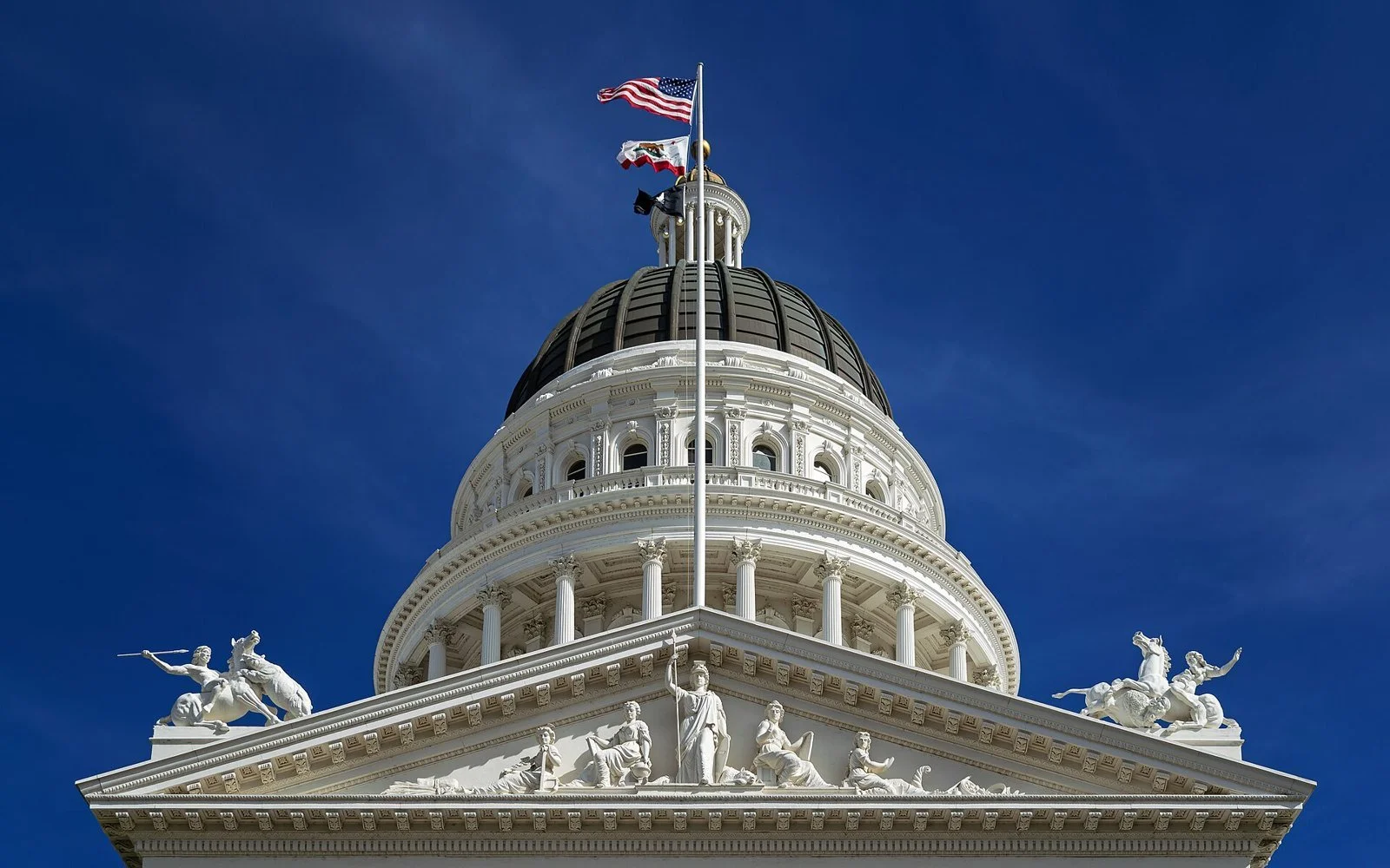Looking up at the white dome, columns and sculpted pediment of the California State Capitol against a clear blue sky.