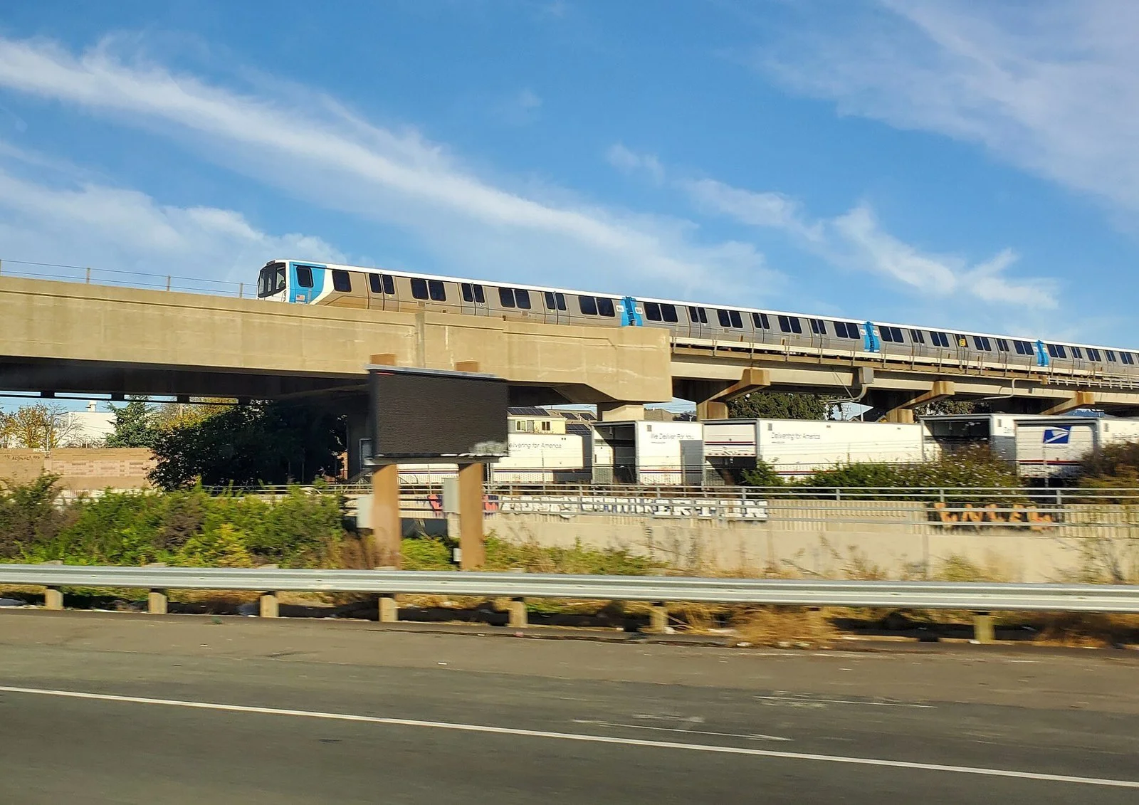 A silver-and-blue BART train runs on an elevated concrete guideway above Interstate 880 in West Oakland under a blue sky with wispy clouds.