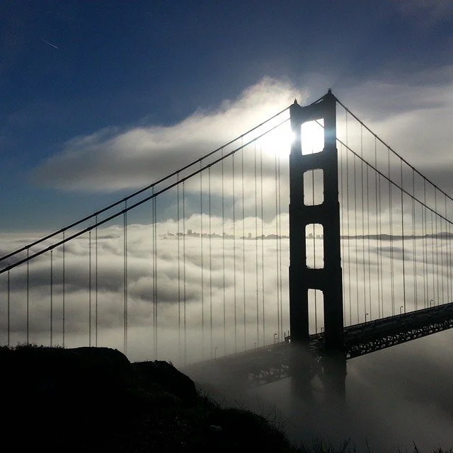 The Golden Gate Bridge spans the strait toward San Francisco, with green coastal hills in the foreground under soft light.