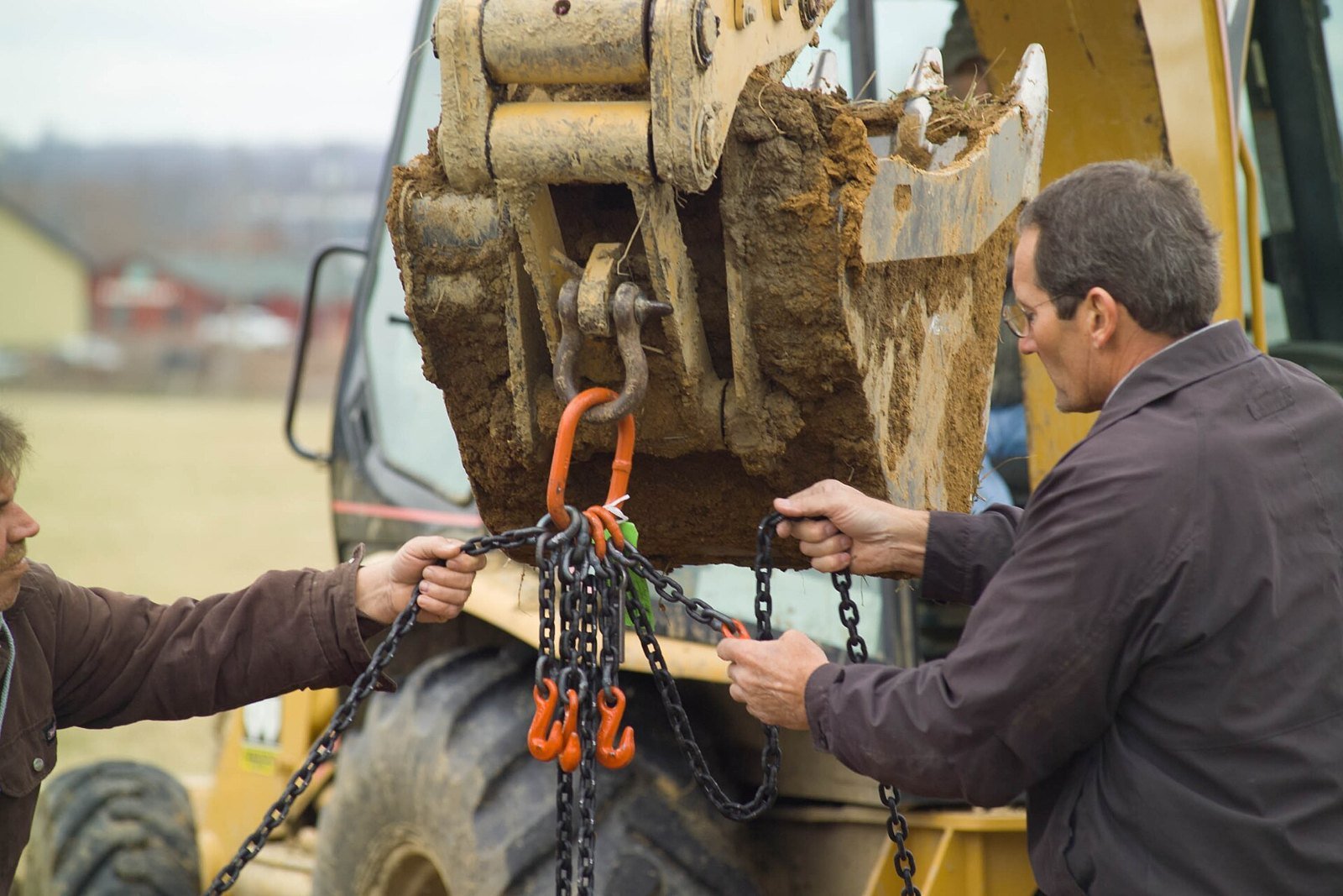 Two construction workers fasten orange lifting chains to the bucket of a yellow backhoe at an excavation site.
