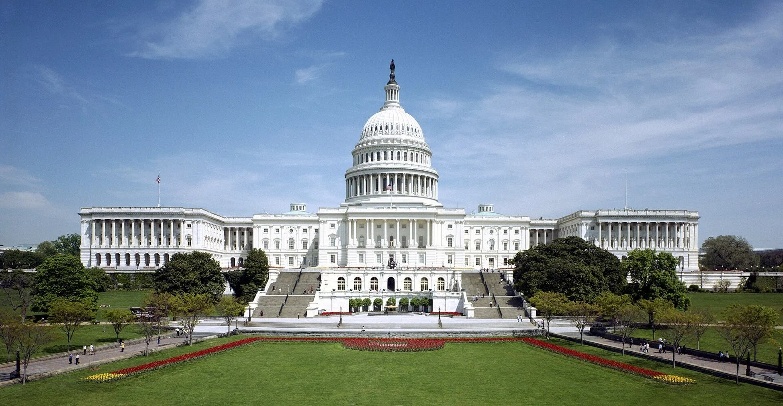The white dome and west facade of the United States Capitol rise above terraces and a reflecting pool under a blue sky.