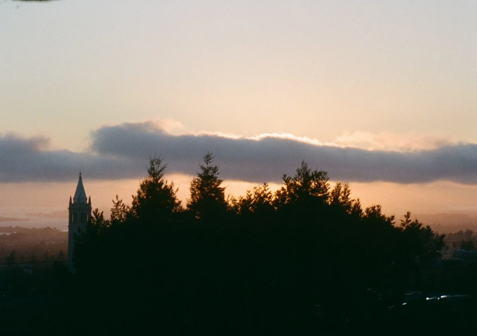 The silhouette of Sather Tower and a line of redwood trees against an orange and blue sunset sky over the Berkeley hills.
