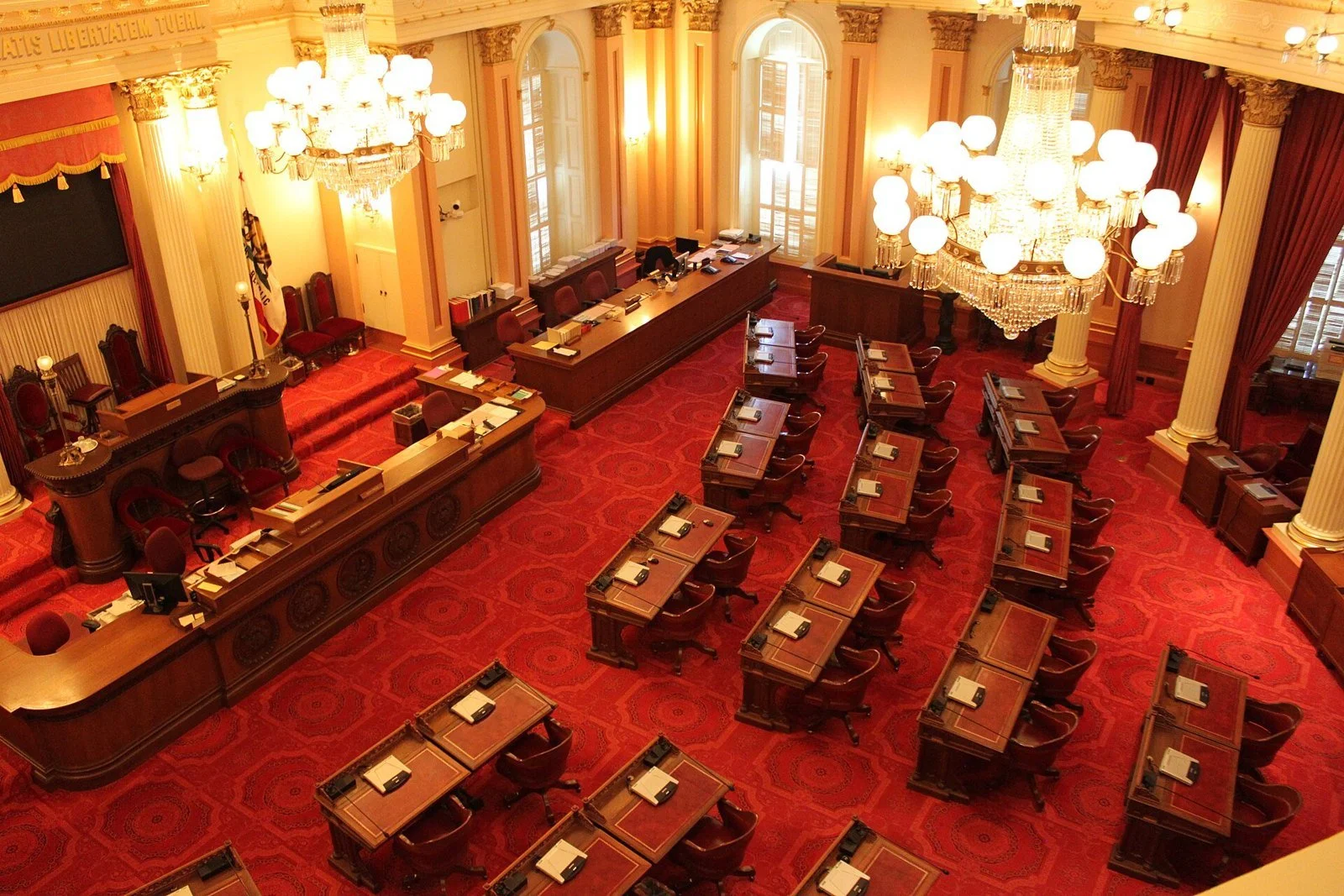 Rows of wooden desks and red carpets fill the California Senate Chamber beneath a coffered ceiling and chandeliers.