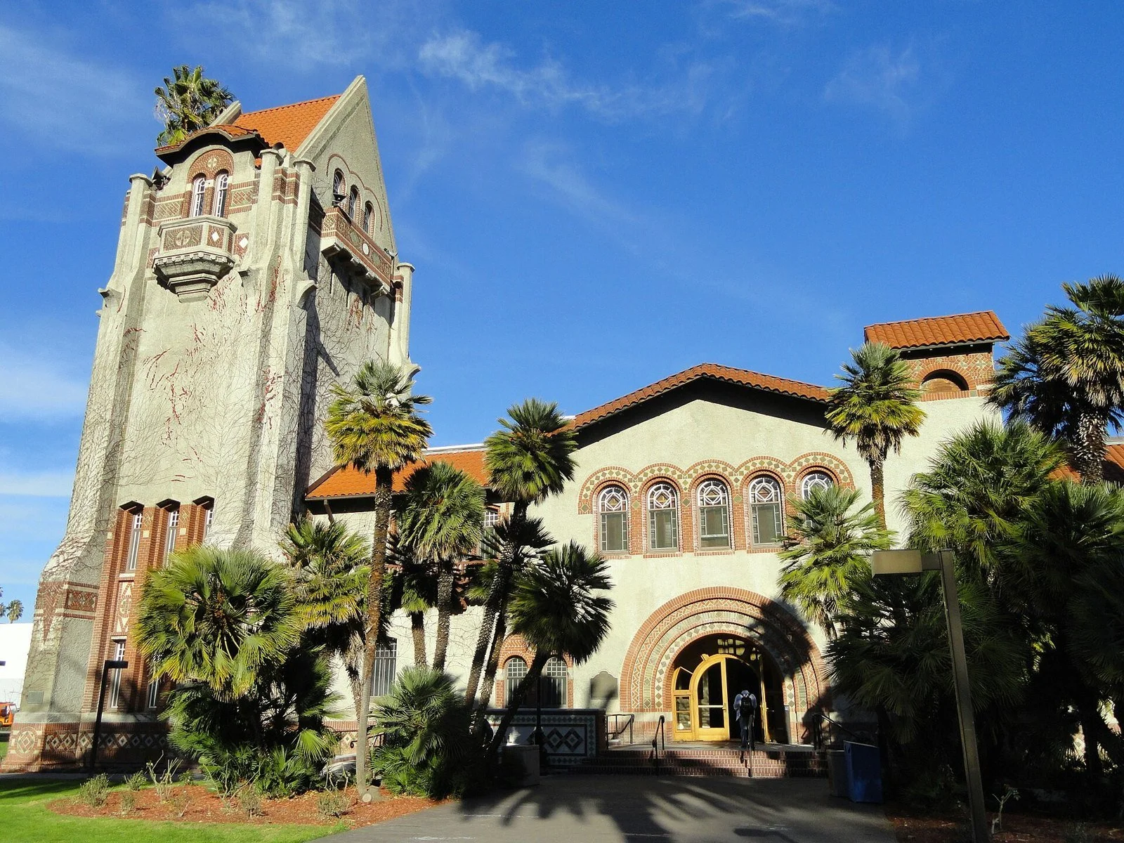 The Spanish Revival brick and stucco Tower Hall at San José State University, with a red-tile roof and palm trees against a blue sky.