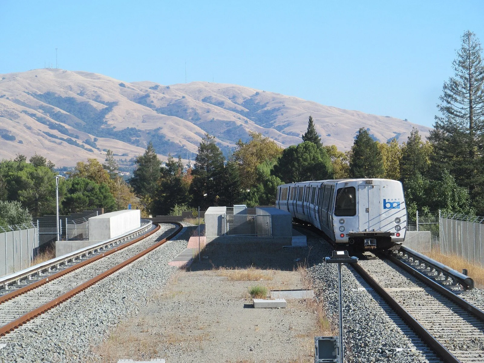A white BART train on elevated tracks heads toward the camera with the brown slopes of Mission Peak and a blue sky behind it.