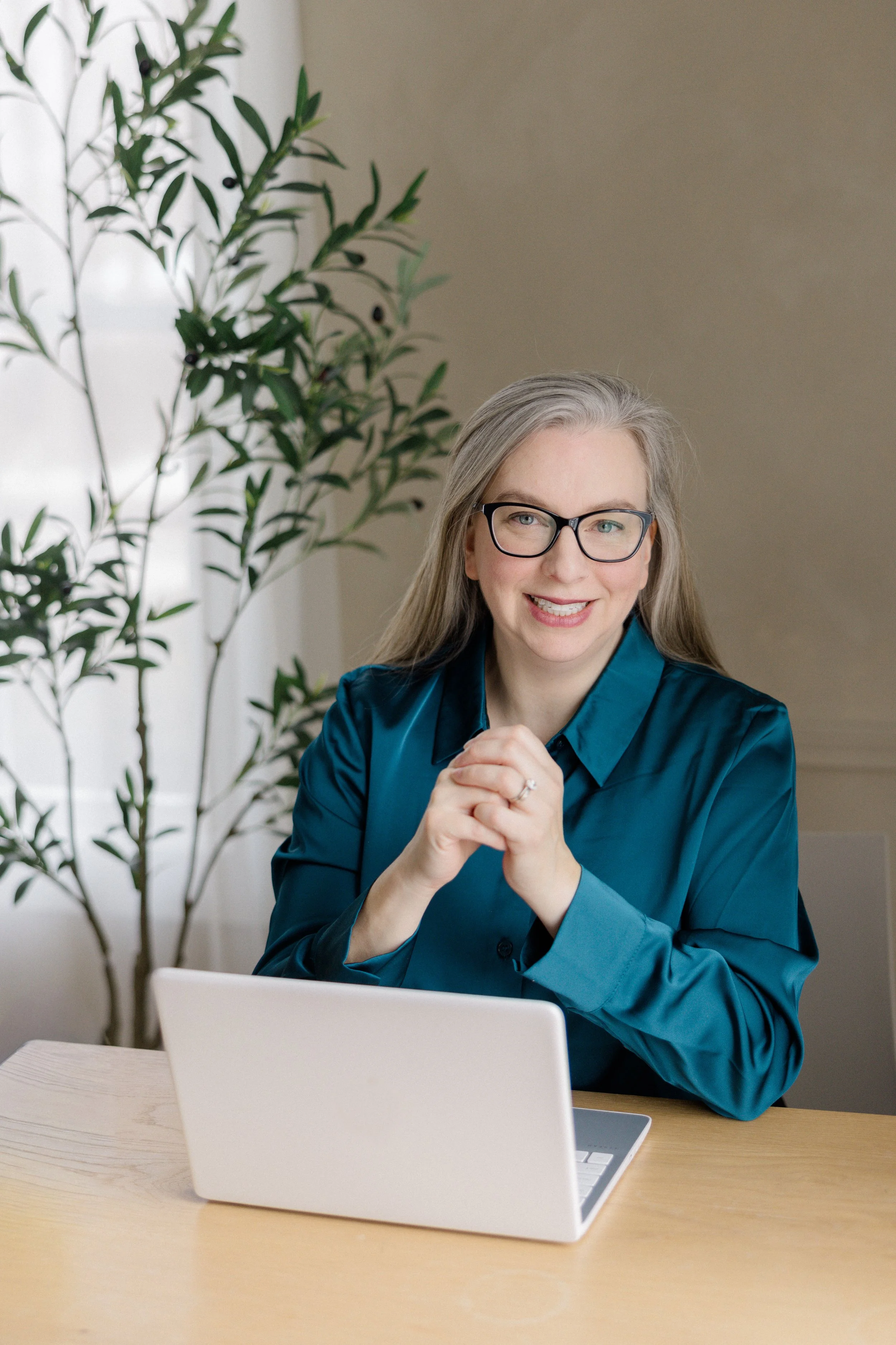 A smiling woman with gray hair and glasses sitting at a light-colored table with a white laptop and tablet, in a room with a large plant and a neutral wall in the background.