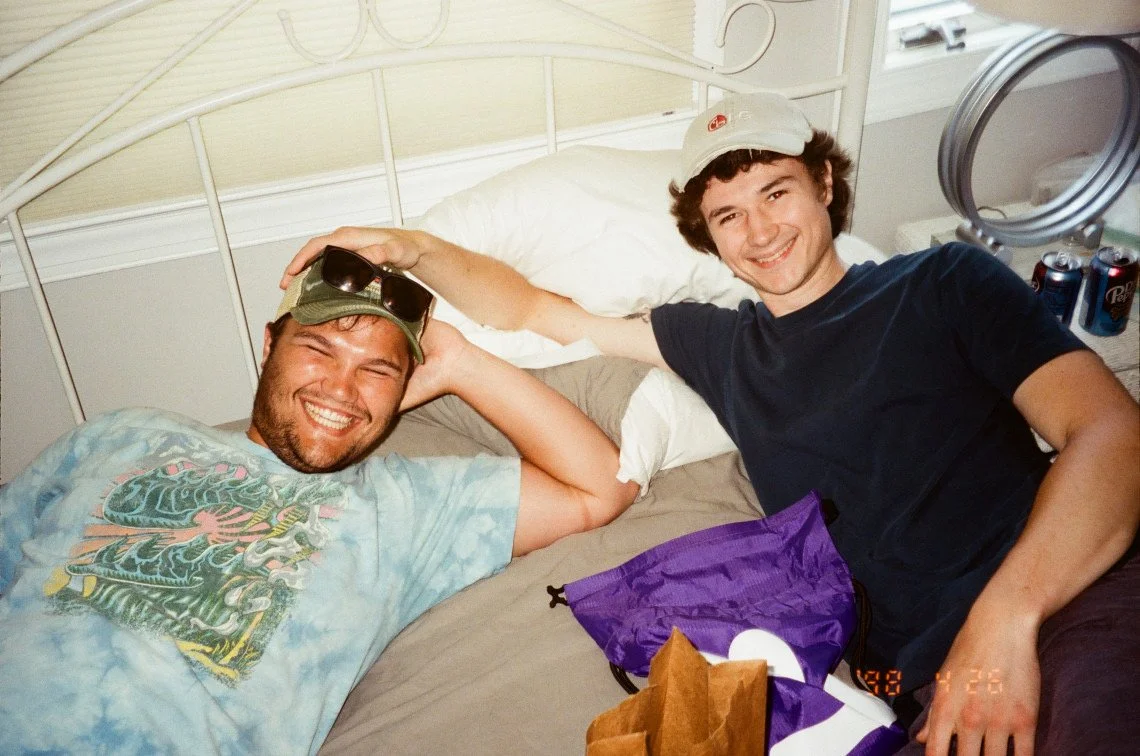 Two young men smiling and relaxing on a bed, one lying down with sunglasses on his head and the other sitting up wearing a baseball cap, with drinks and snacks nearby.