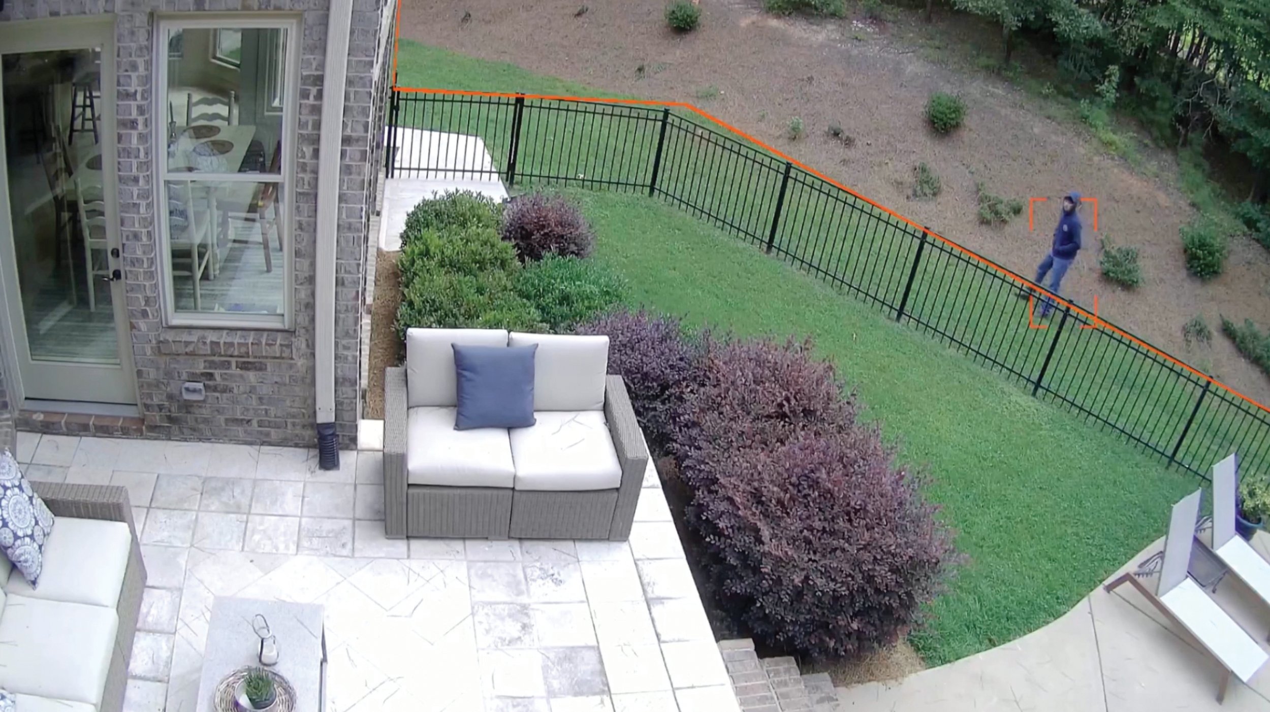 View of a backyard with a patio, outdoor furniture, green lawn, and a black metal fence. A person in a jacket and jeans is standing near a garden area with bushes, trees, and a dirt slope.