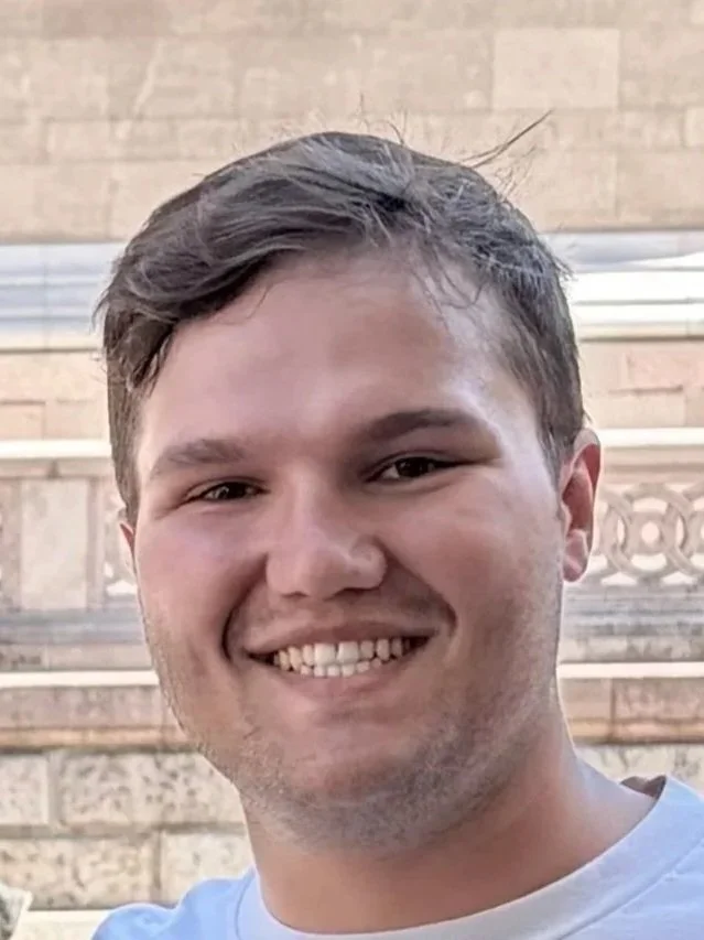 Close-up of a young man smiling outdoors, with a brick building and metal railing in the background.