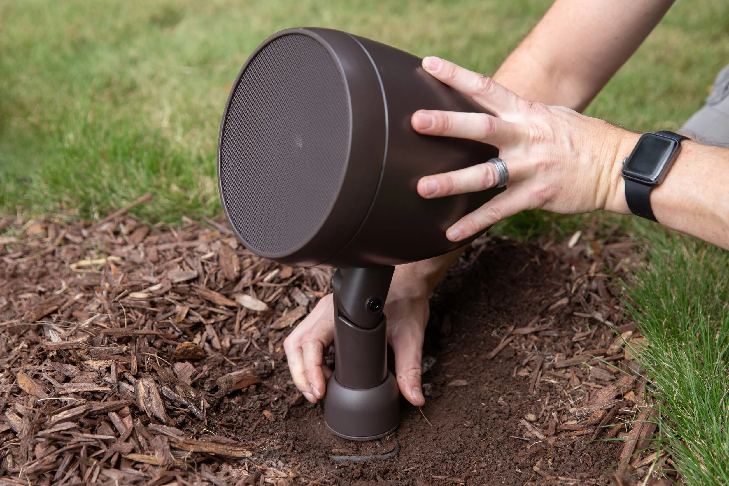 Person installing a black portable speaker into the ground with their hands during outdoor activity.