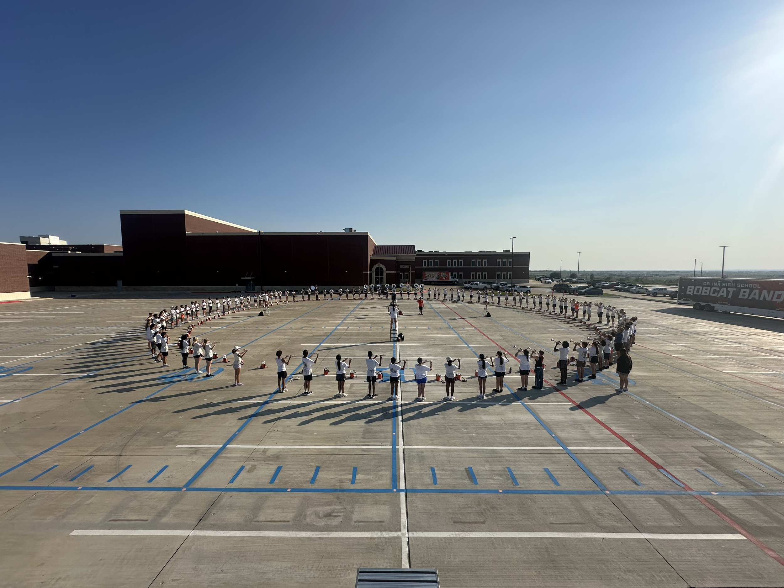 Students in uniform gathered in a large outdoor parking lot, practicing a drill or formation, with a school building and parking lot in the background during daytime.