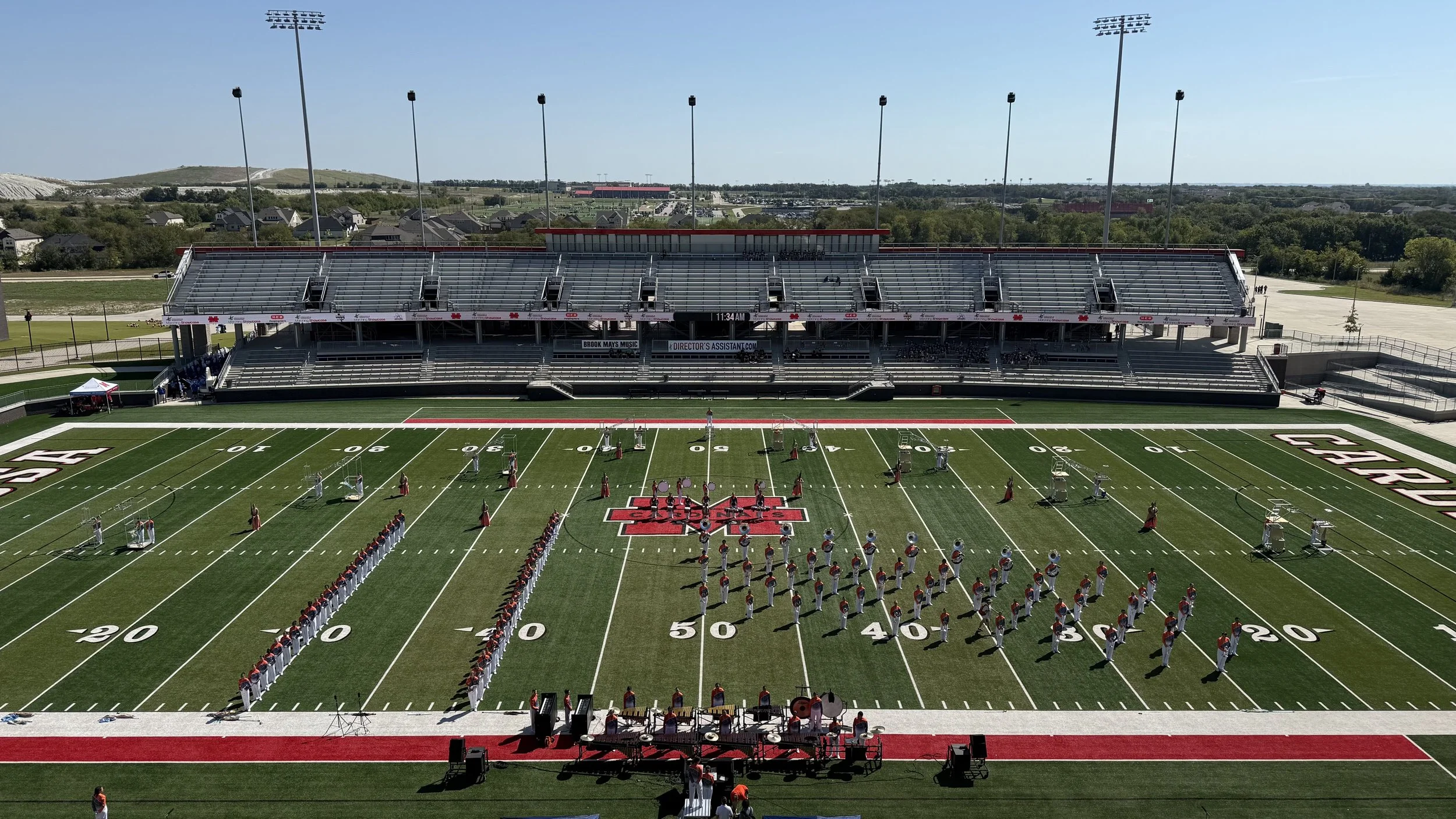 A marching band performing on a football field with stadium bleachers in the background. The football field has yard lines and a large red and white logo at the center. The band members are dressed in red, white, and blue uniforms, and there is a conductor at the front. Tall light poles are visible around the field, and a small tent is set up on the sidelines.