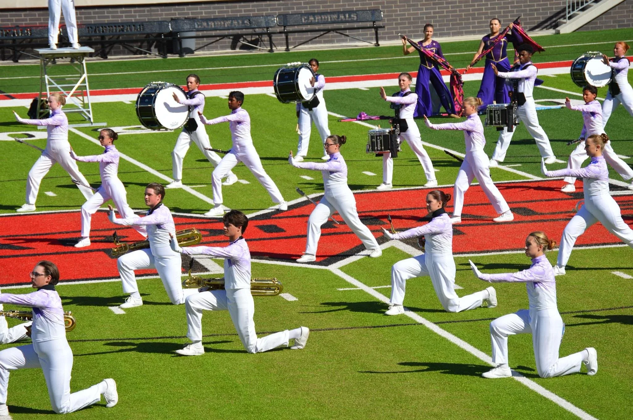 A marching band performance on a football field with drummers, baton twirlers, and musicians in white uniforms with purple accessories.
