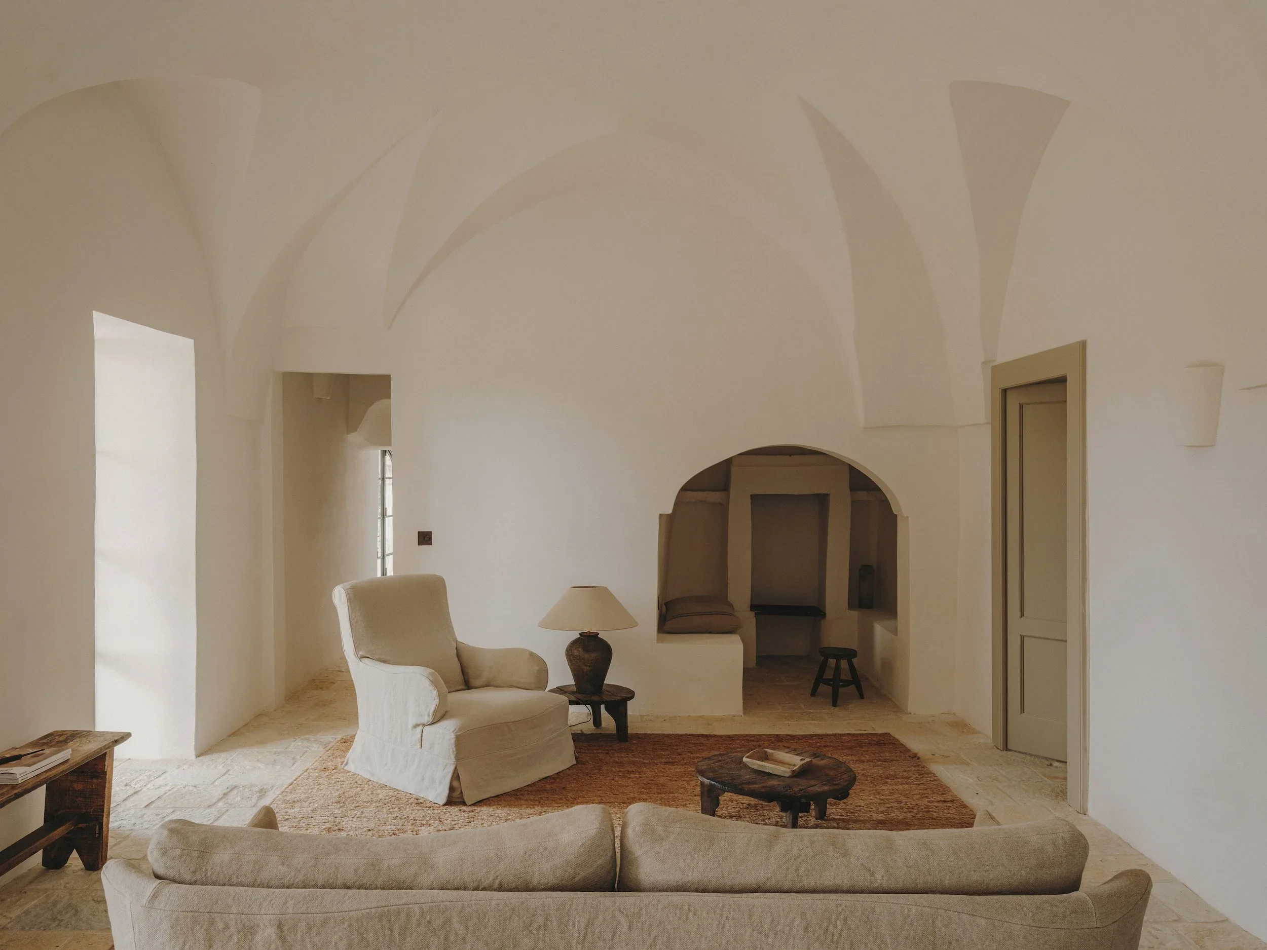 Living room with beige armchair, beige sofa, wooden side table, lamp, and small round wood coffee table on a terracotta rug. Arched ceiling and small alcove in the background.