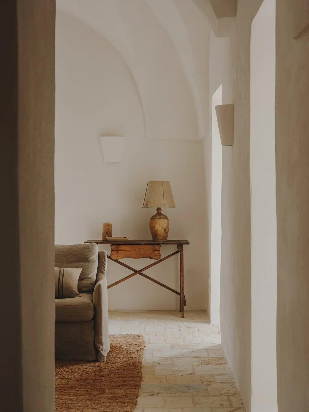Living room with a beige sofa, a wooden console table with a textured ceramic table lamp, a small decorative item, and wall-mounted light fixtures, with textured walls and stone flooring.