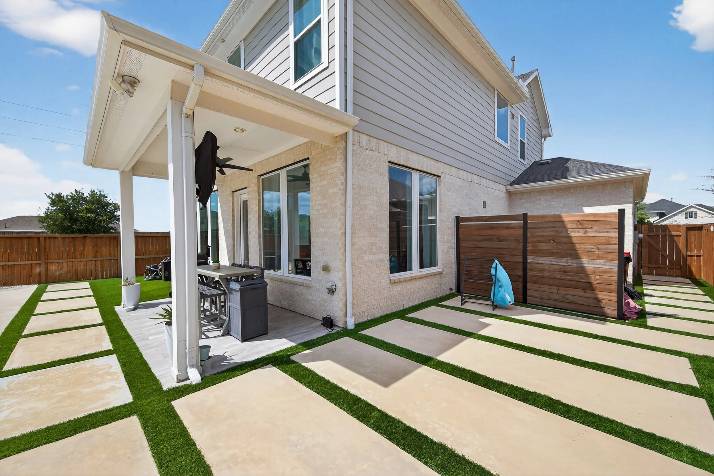 Backyard patio area with large concrete pavers separated by grass strips, outdoor dining table under covered porch, wooden privacy fence, and neighboring houses in the background.