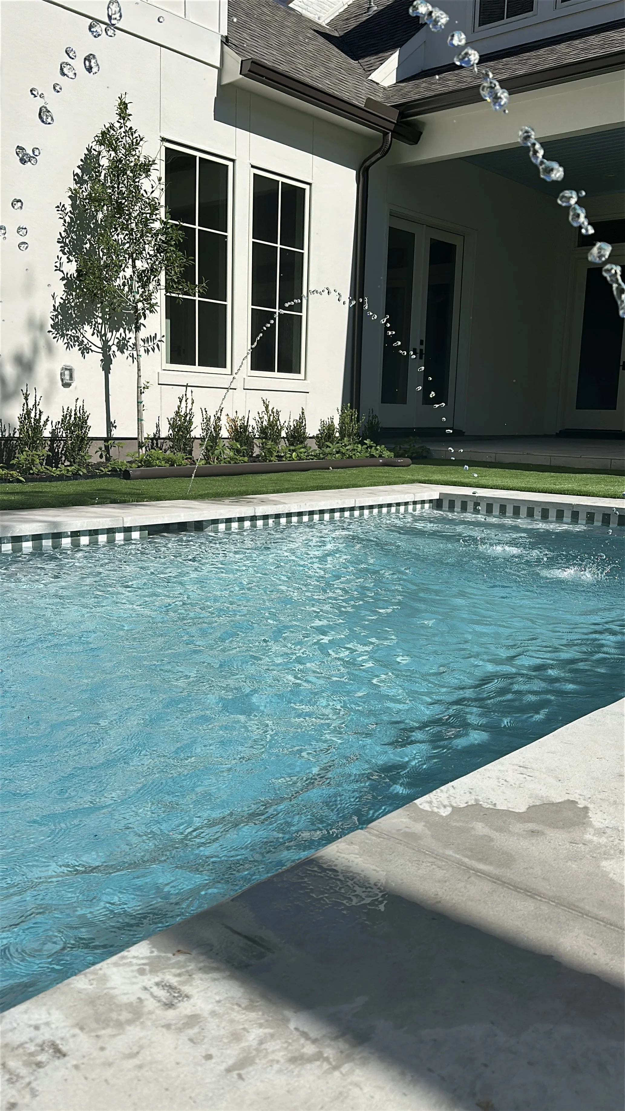 A backyard swimming pool with water splashing from a fountain in a fenced backyard, with a modern house exterior featuring large windows, a patio door, and a small tree planted near the house wall.