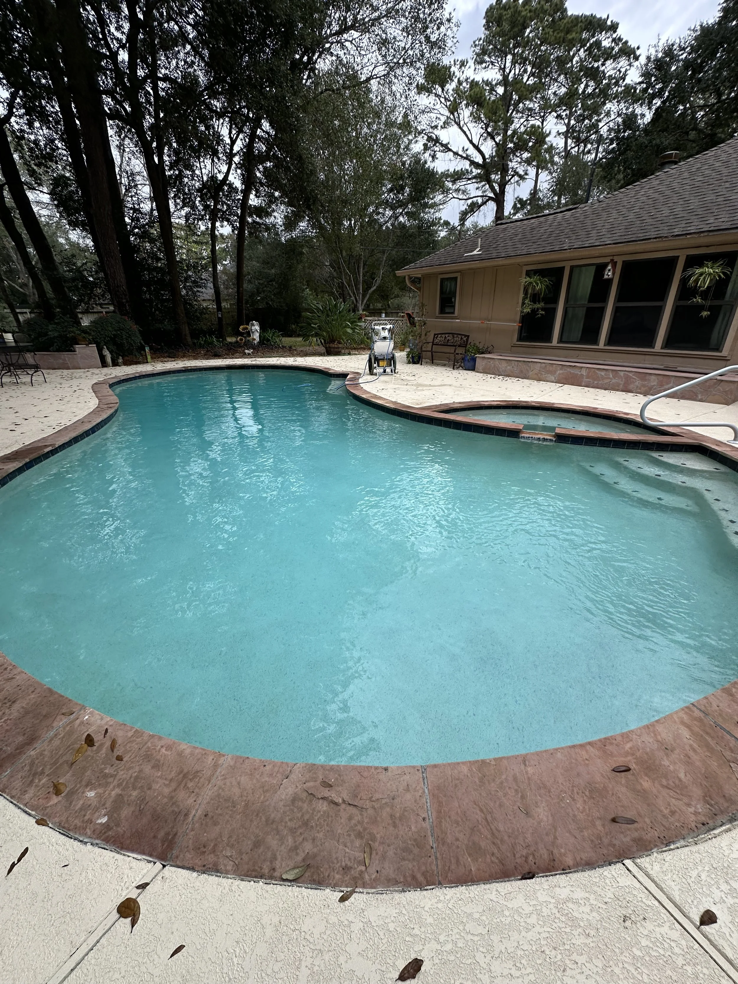 A backyard swimming pool with a curved shape, surrounded by a concrete deck with some leaves scattered on it. There is a hot tub adjacent to the pool, and the area is bordered by trees and a house with windows and a sloped roof.