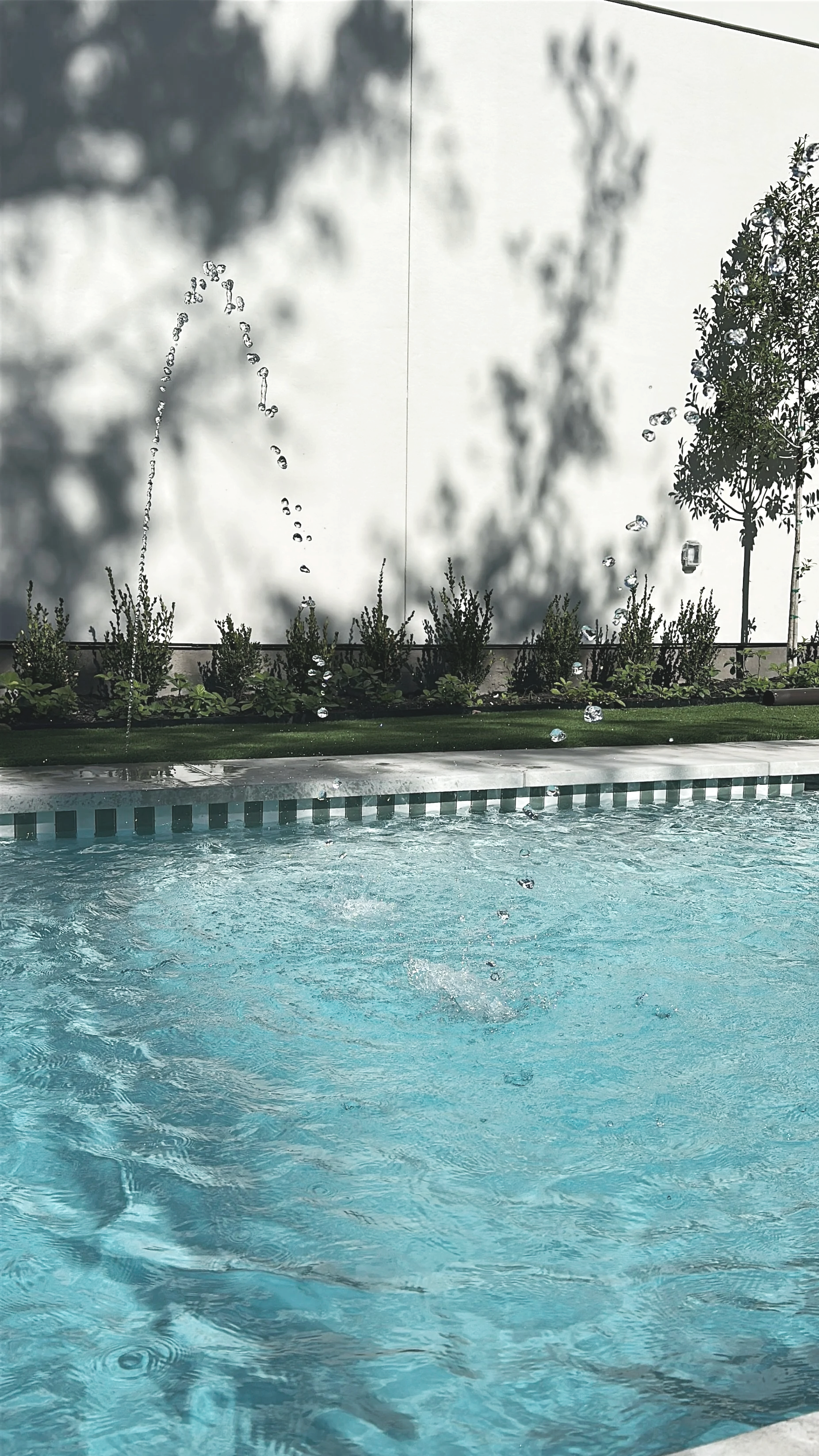 Swimming pool with water splashing from a fountain, greenery along the edge, and shadows of trees and a wire overhead on a sunny day.
