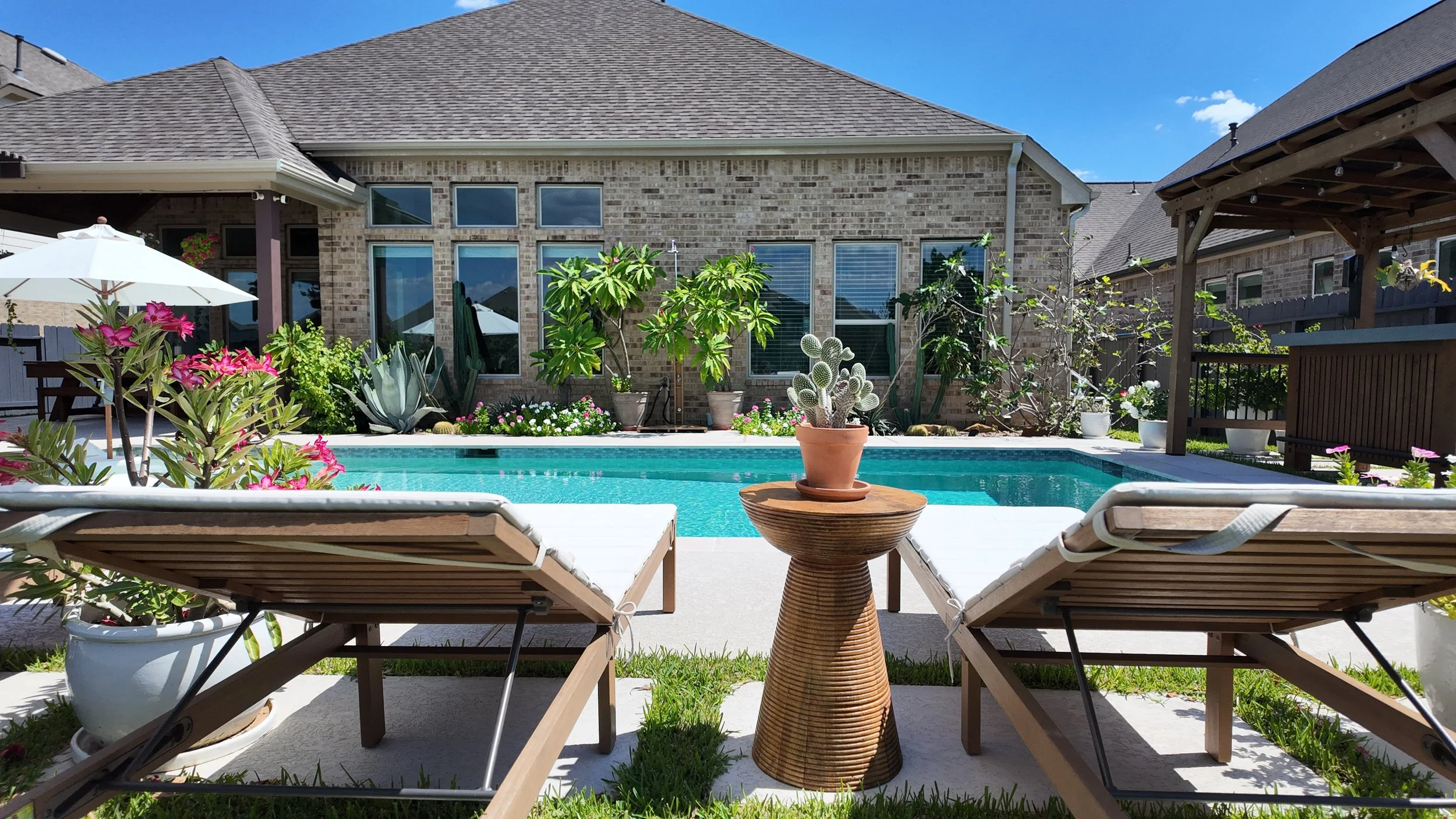 Backyard with a swimming pool, two lounge chairs, potted plants, and a patio, with a brick house and a blue sky in the background.