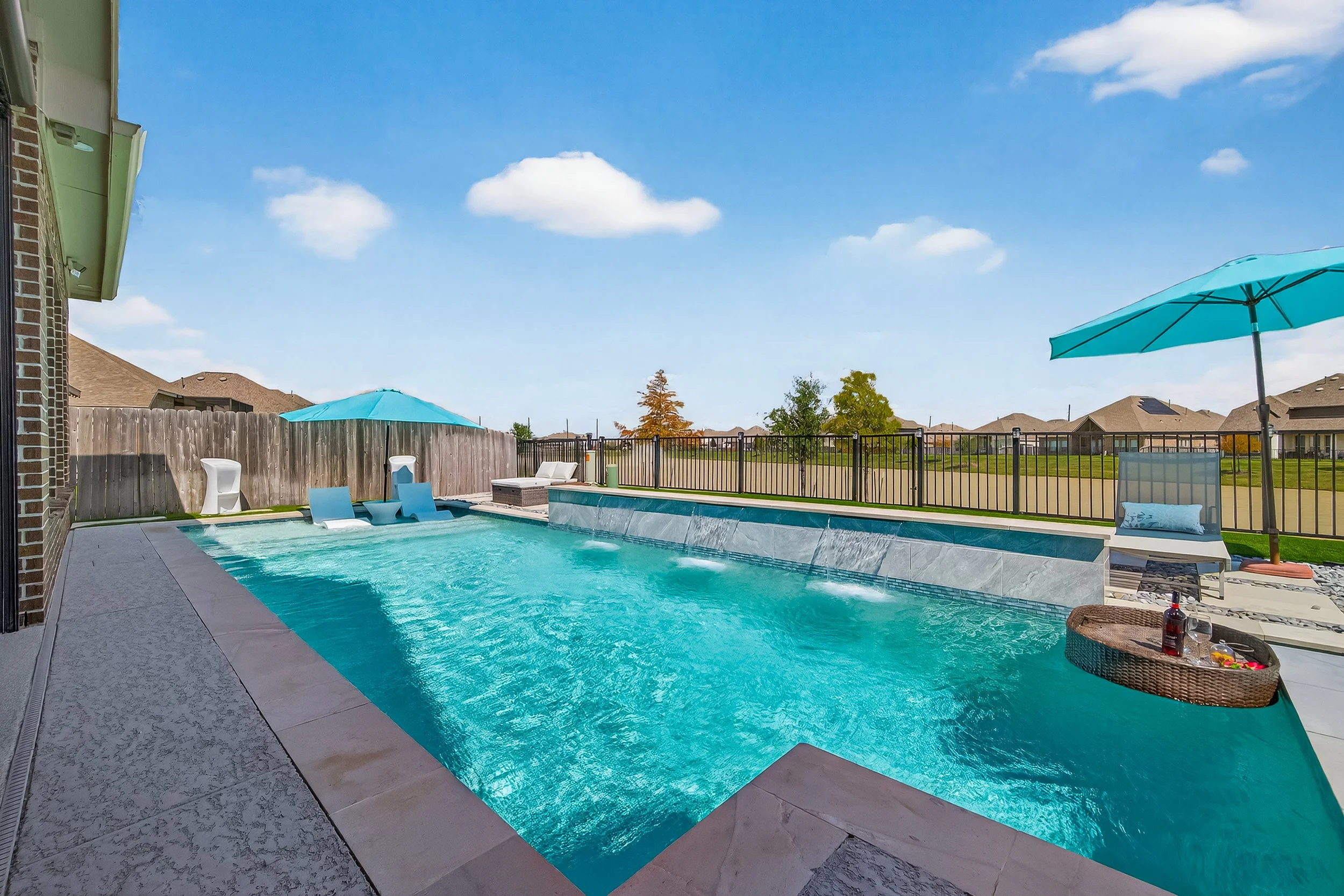 Backyard swimming pool with water feature, poolside chairs, umbrellas, and a wicker tray with drinks and snacks under a clear blue sky.