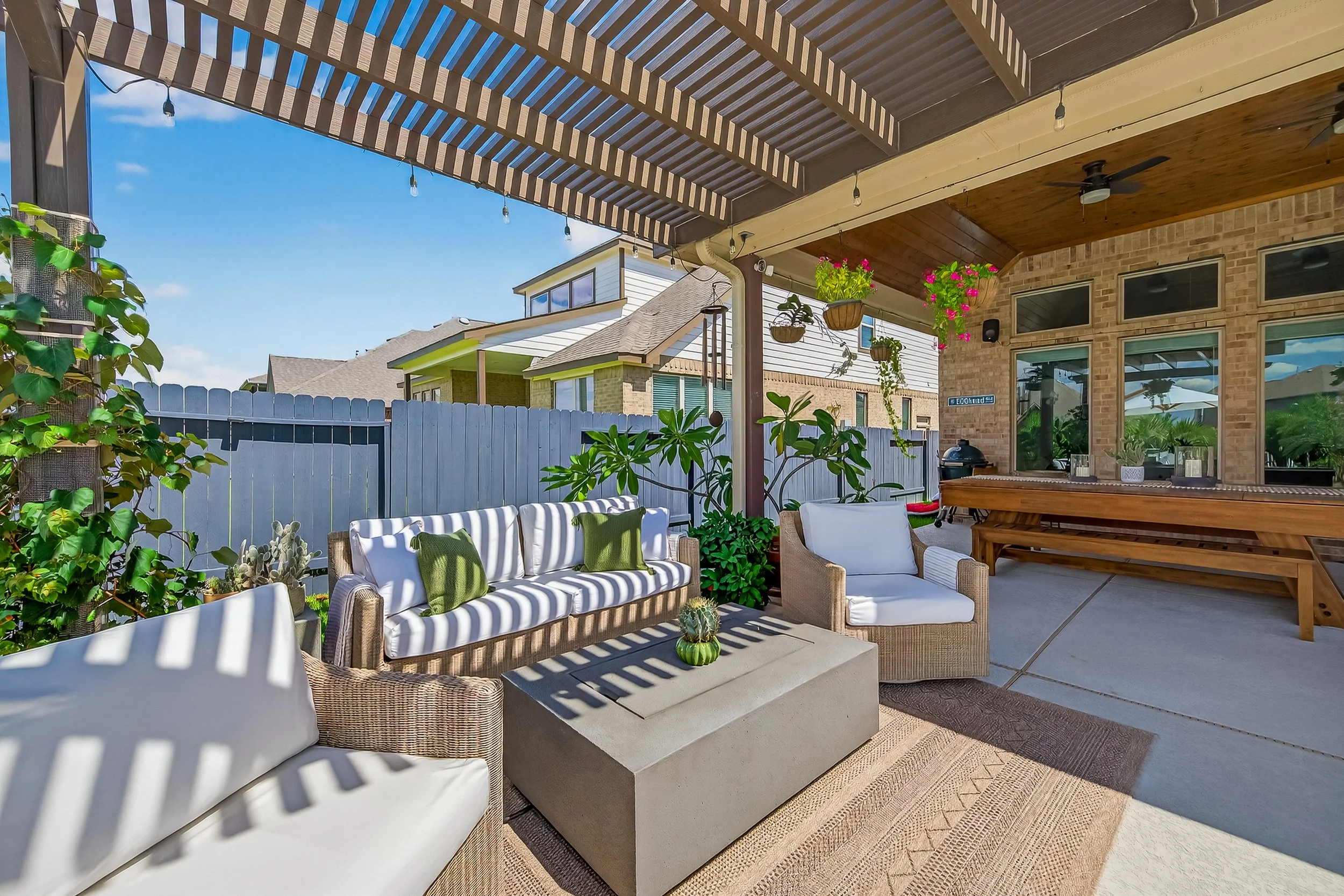 Outdoor patio with wicker and cushioned seating, potted plants, hanging flower baskets, a concrete coffee table with decorative cactus, a wooden pool table, and a fenced backyard with neighboring houses under a blue sky.