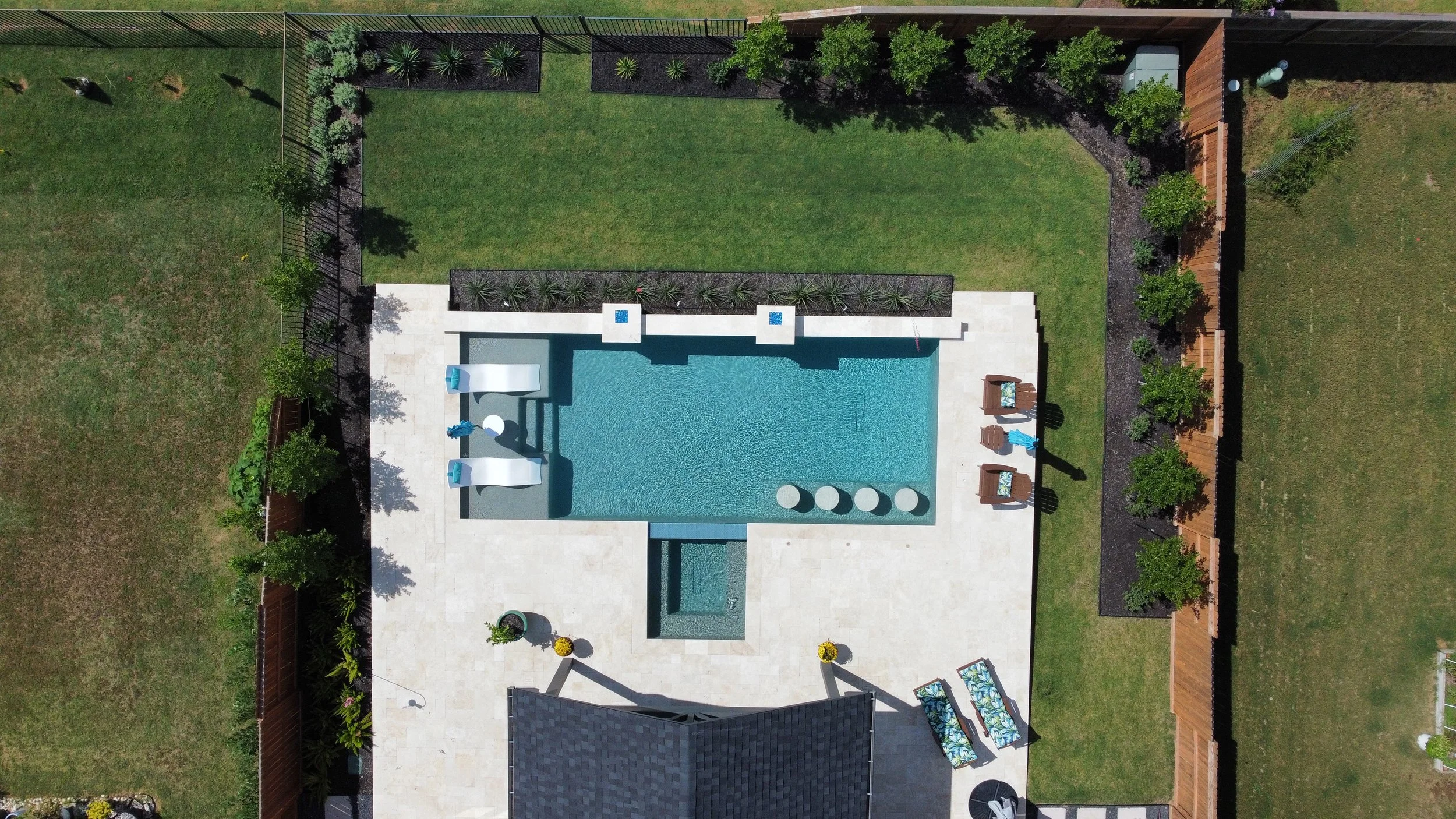 Aerial view of a backyard featuring a rectangular swimming pool with a hot tub in the lower center, surrounded by a light-colored patio with lounge chairs, umbrellas, and ornamental plants, enclosed by a wooden fence with green trees and landscaped garden areas.