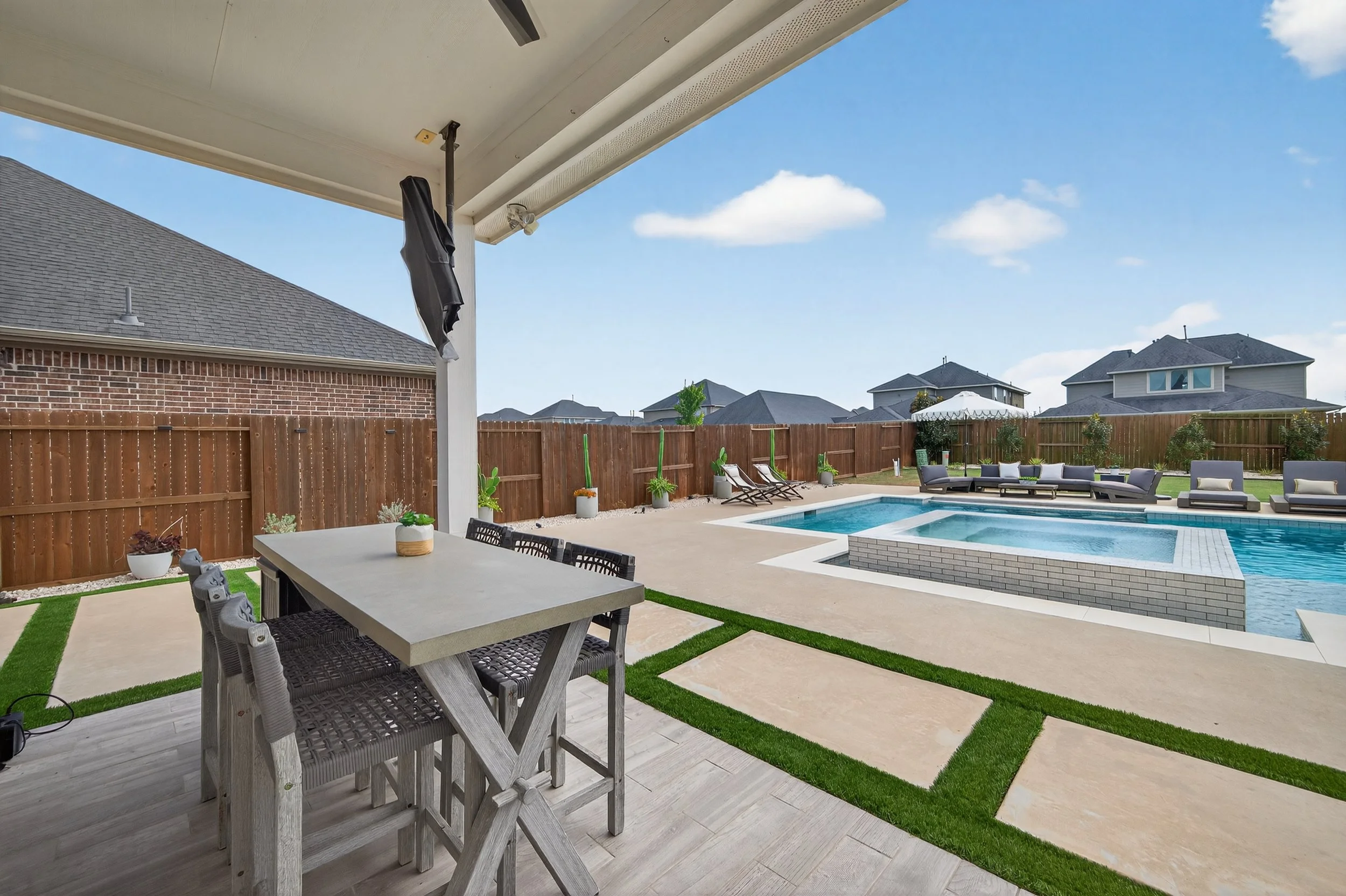 A backyard with a covered patio, a rectangular swimming pool, lounge chairs, and outdoor seating, surrounded by a wooden fence under a blue sky with a few clouds.