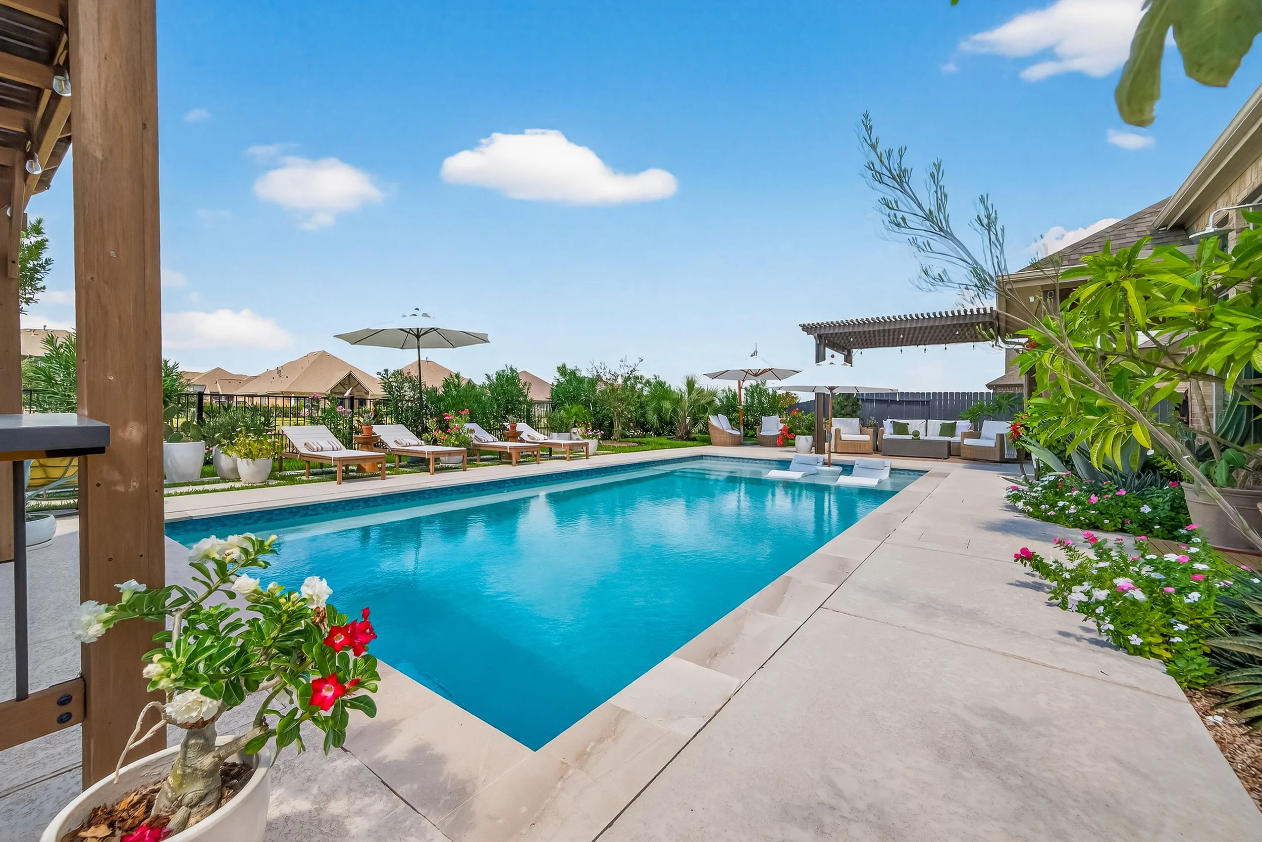 A backyard swimming pool area with lounge chairs, umbrellas, potted plants, and outdoor seating on a sunny day.