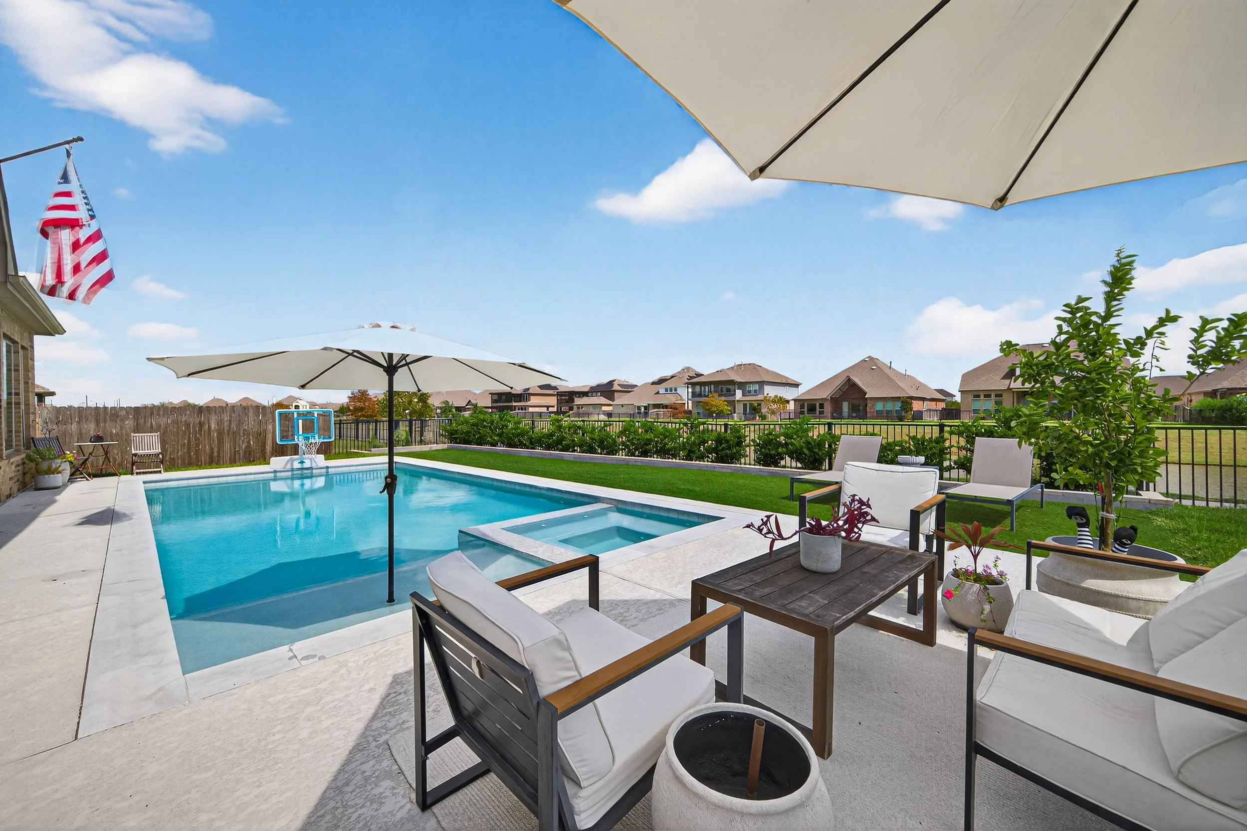 Backyard with swimming pool, patio furniture, umbrellas, and a fence, with houses and clear blue sky in the background.
